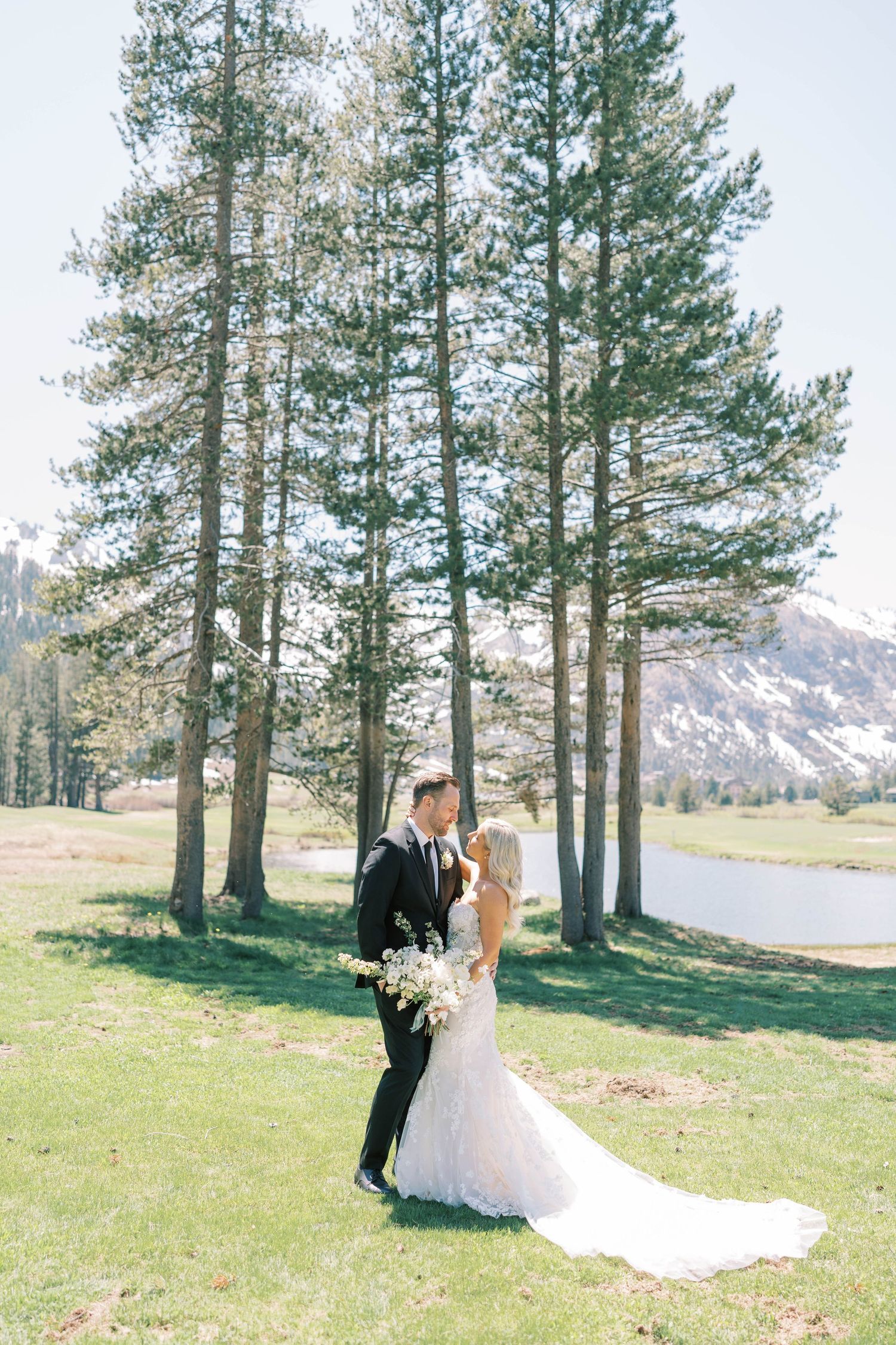 Newlyweds stand together in an outdoor mountain setting with evergreen trees and snow-capped peaks.