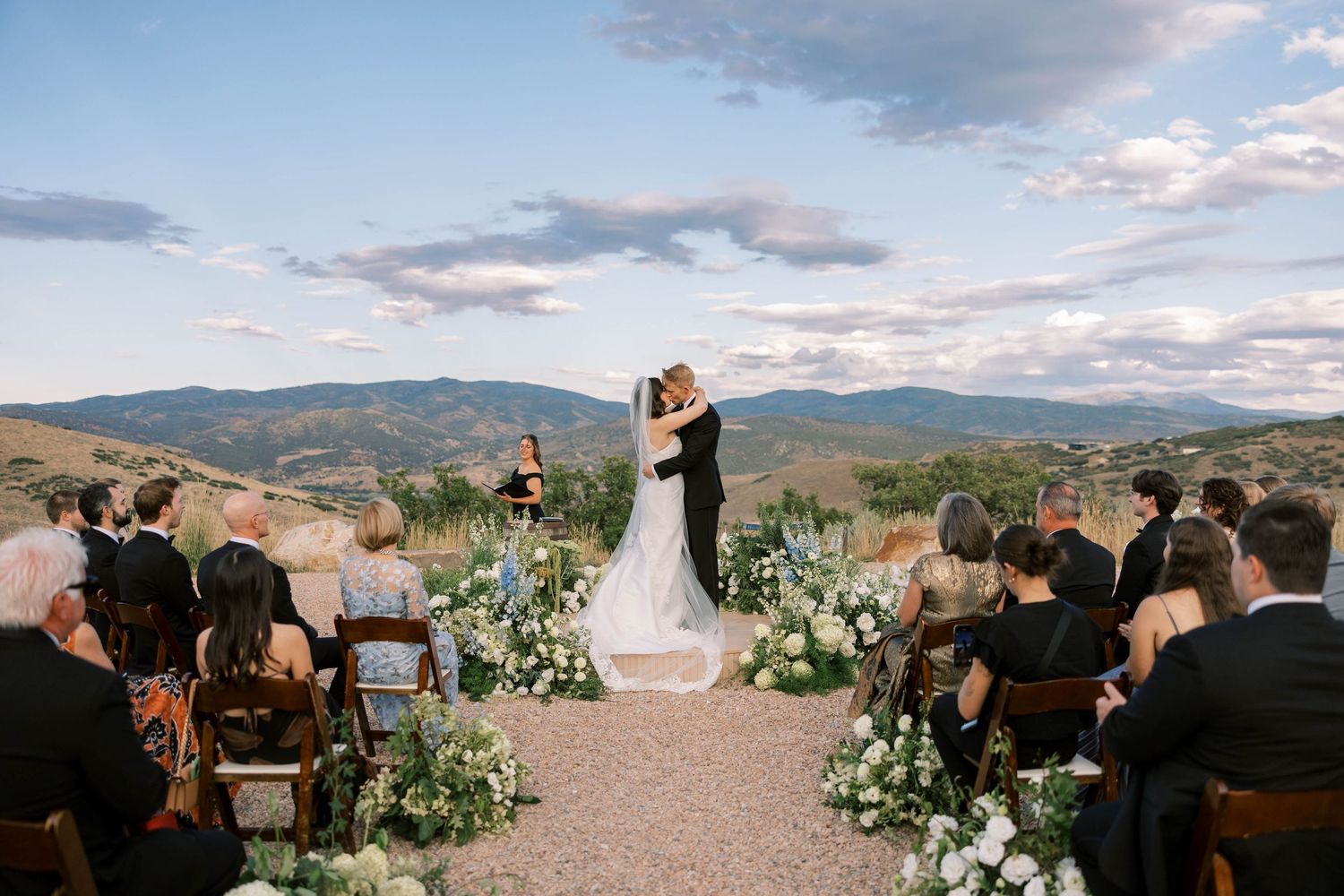 A panoramic view of an outdoor wedding ceremony in the mountains with floral arrangements and guests seated on wooden chairs.