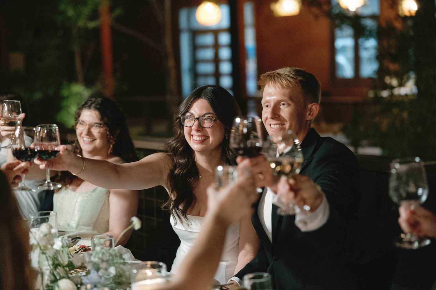 Wedding reception celebration with guests raising glasses for a toast in dimly lit venue.