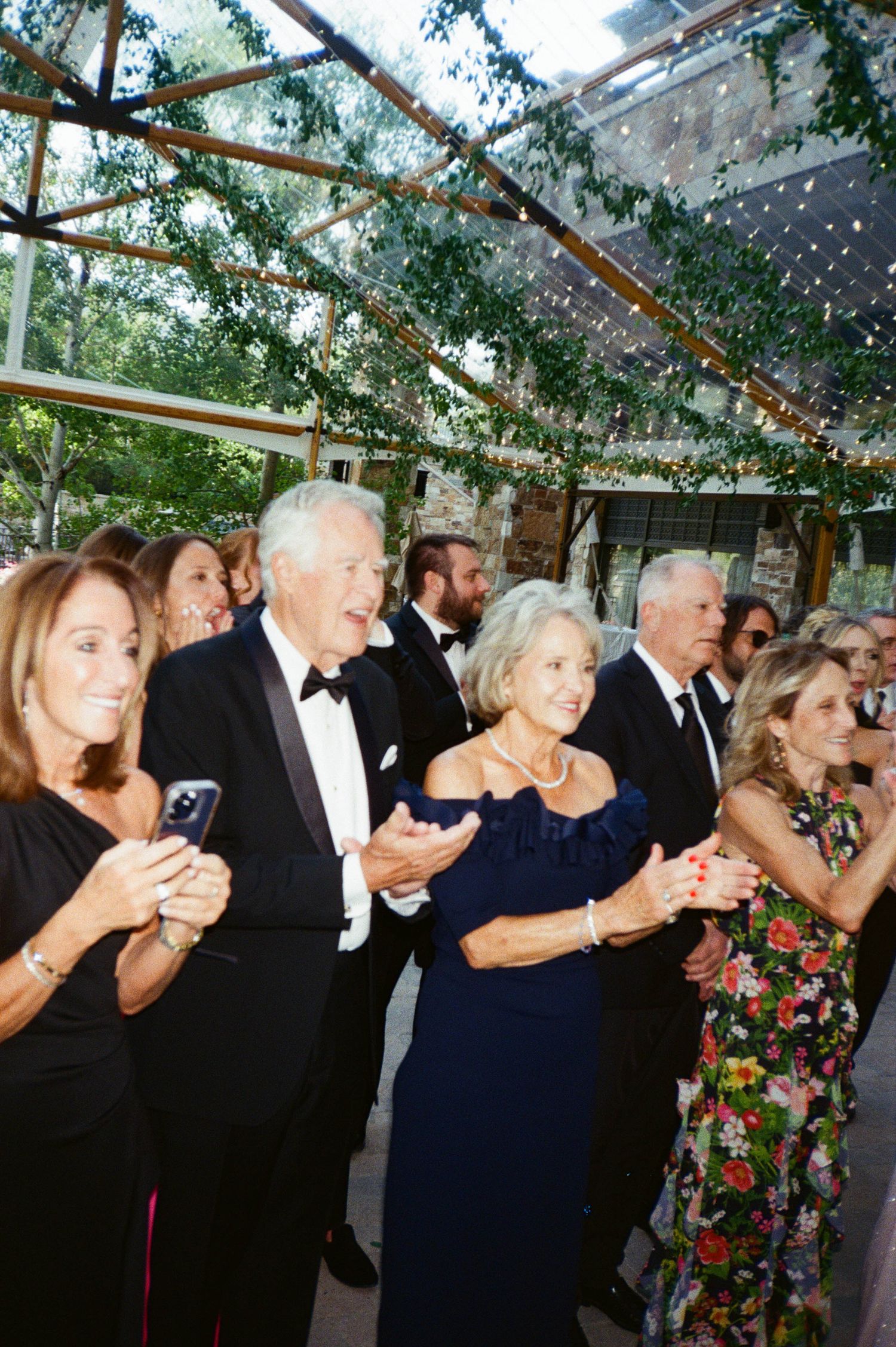 Wedding guests in formal attire dancing together under a clear tent with string lights and floral decorations.