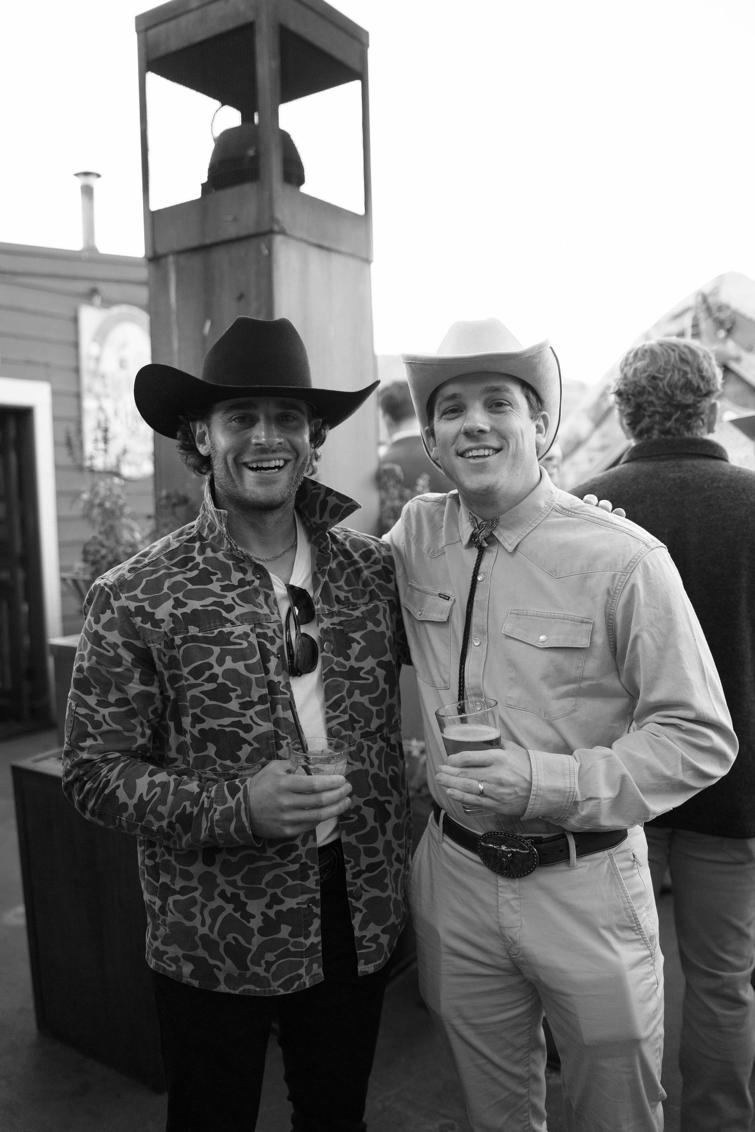 Two men in western attire with cowboy hats smile while holding drinks at an outdoor event.