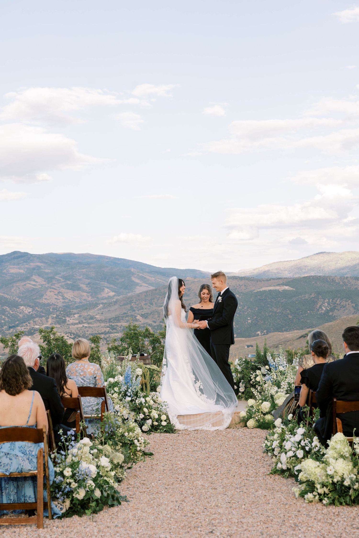 Outdoor mountain wedding ceremony with white floral arrangements and wooden chairs against a scenic backdrop.