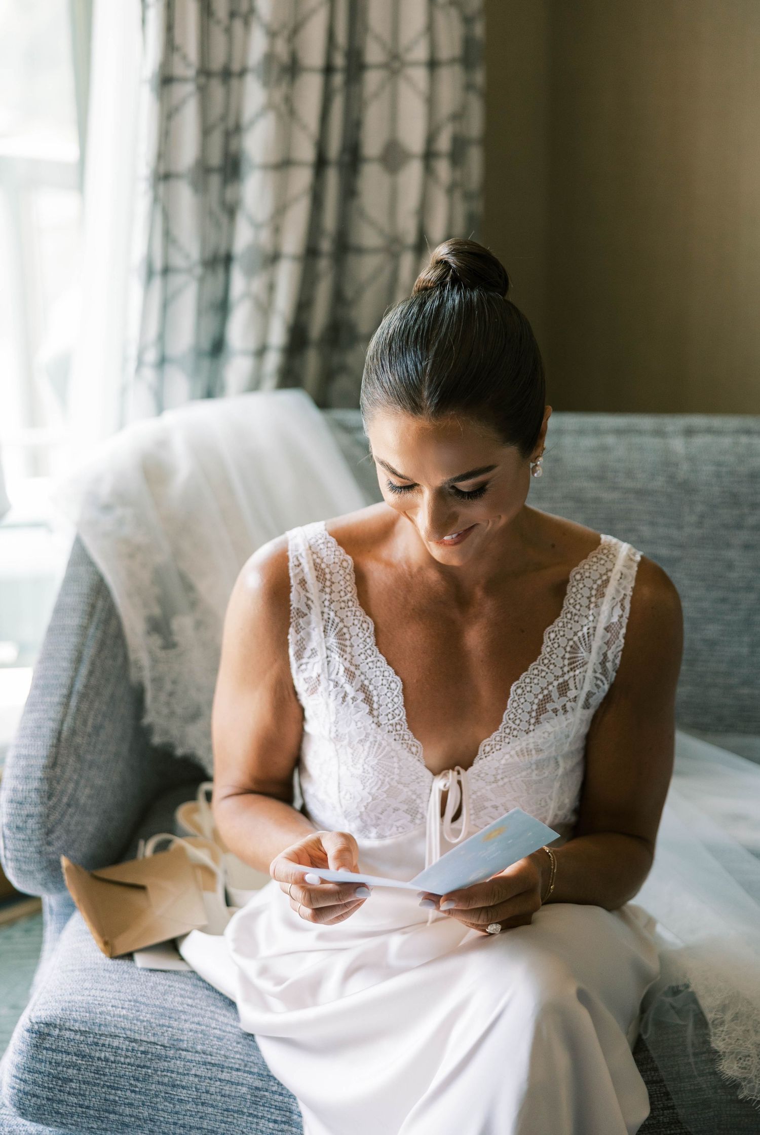 A bride in a white lace dress sits on a gray couch while writing in a journal or notebook.