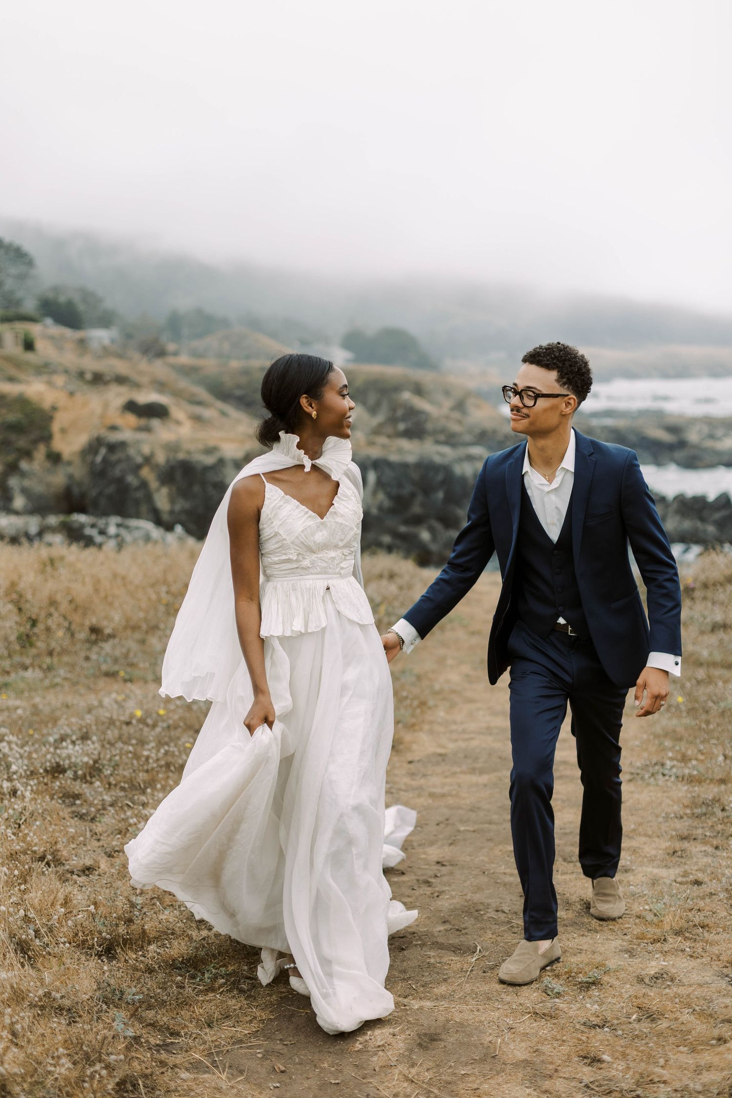 A couple in wedding attire playfully runs and dances together on a misty coastal cliff during their outdoor celebration.