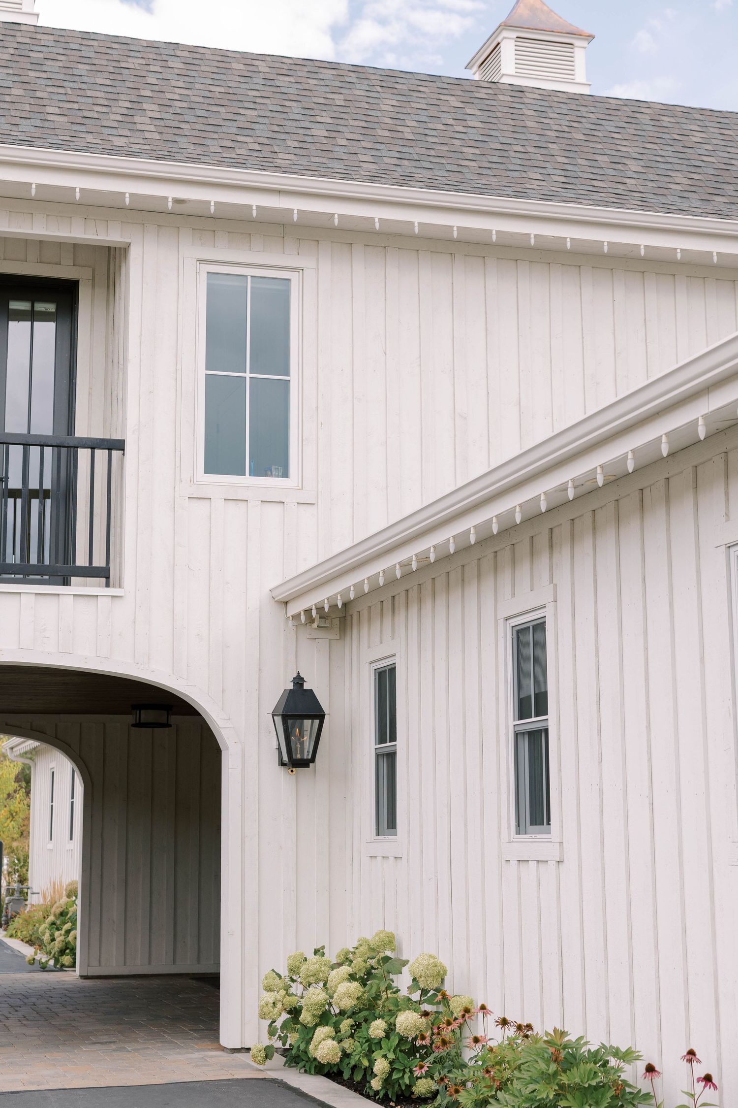 White barn-style building with vertical siding features an arched entryway and black lantern beside blooming hydrangeas.