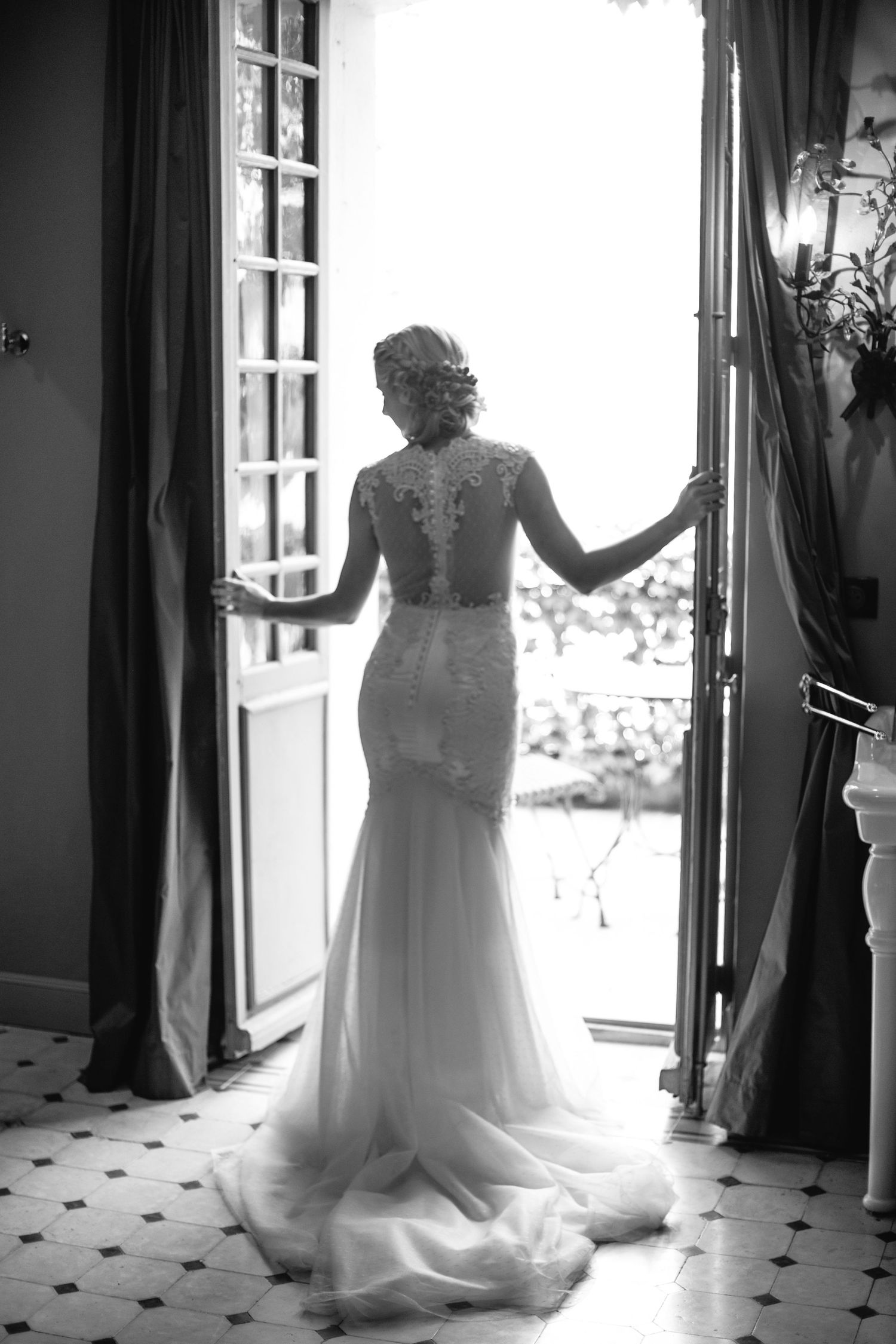 Silhouette of a bride in lace gown standing in doorway against bright window light.