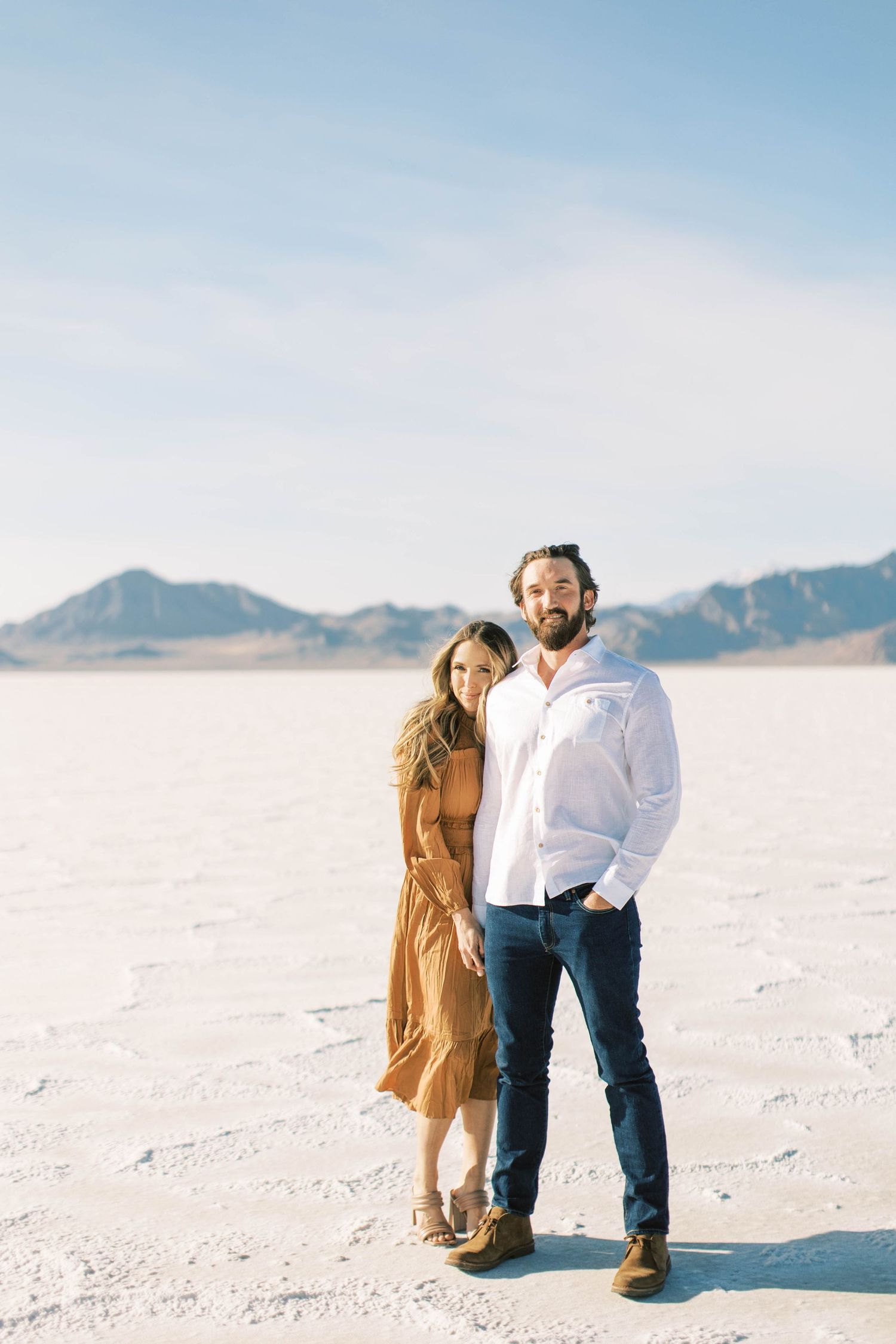 Couple in casual attire posing together on salt flats with mountain range in background.