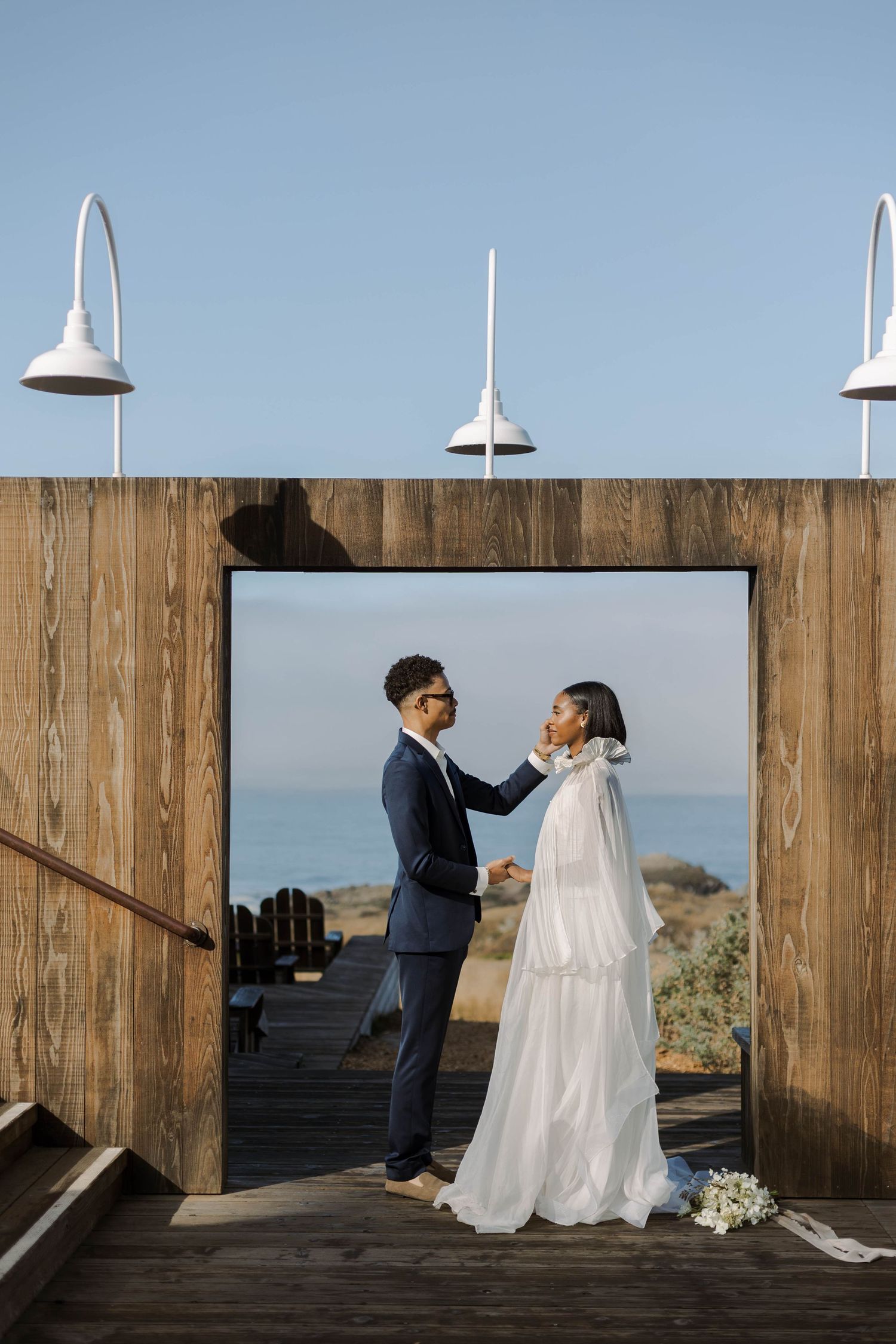 A couple shares a playful moment with a wedding dress on a wooden deck overlooking the ocean at sunset.