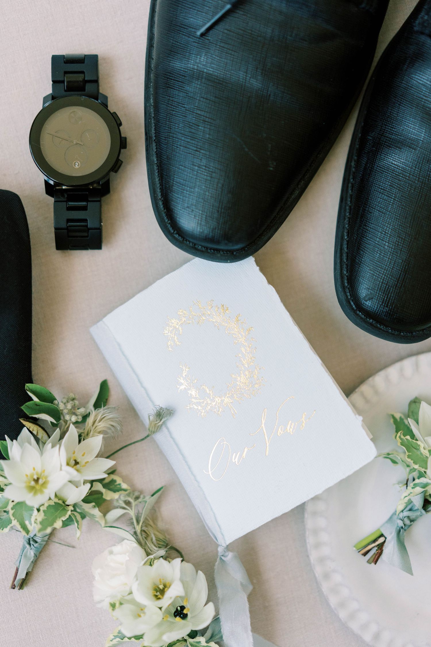 Wedding details including black dress shoes, watch, and white vow book with floral accents.