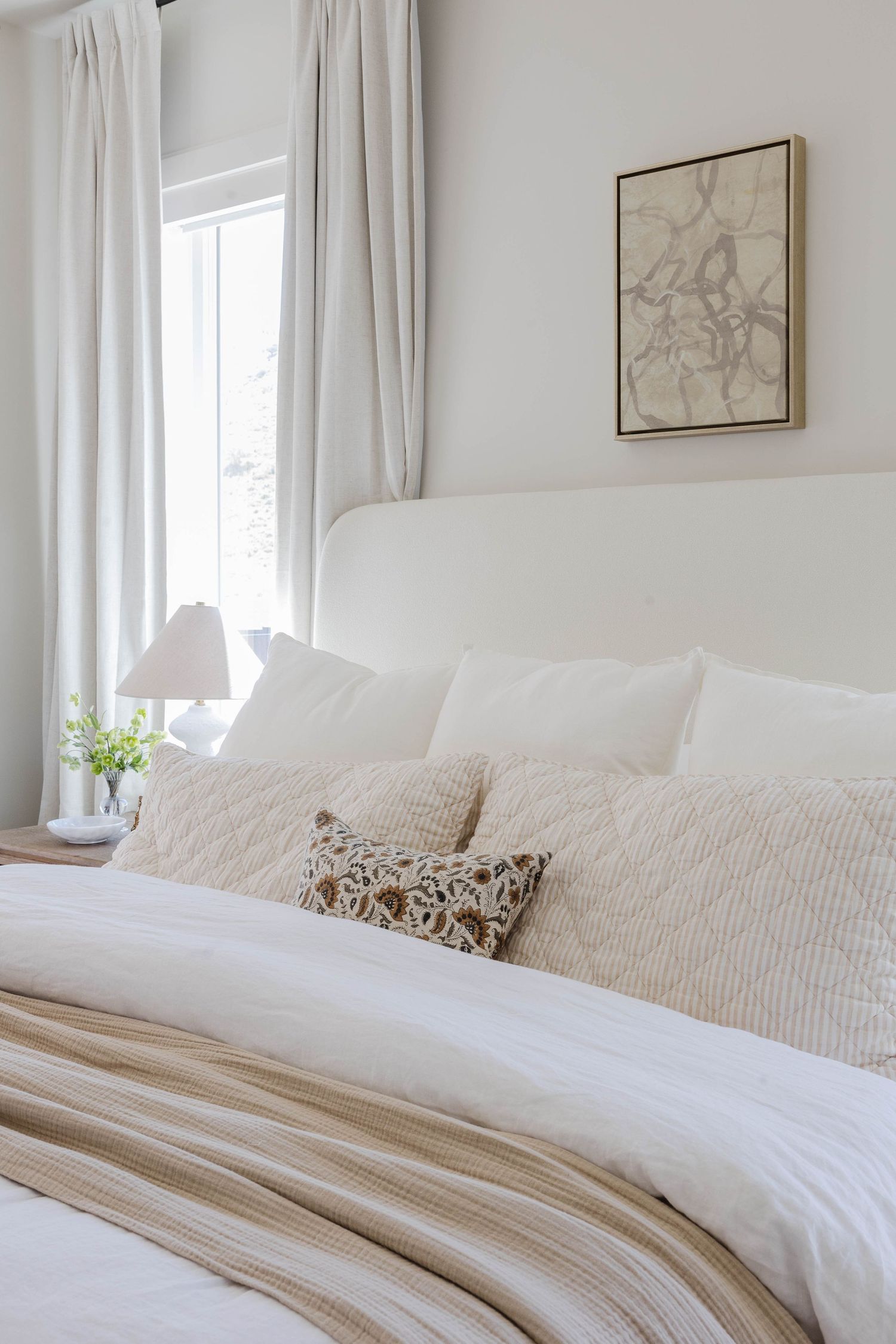 Serene white bedroom with upholstered headboard, layered neutral pillows, linen throw, and soft natural light from curtained window.