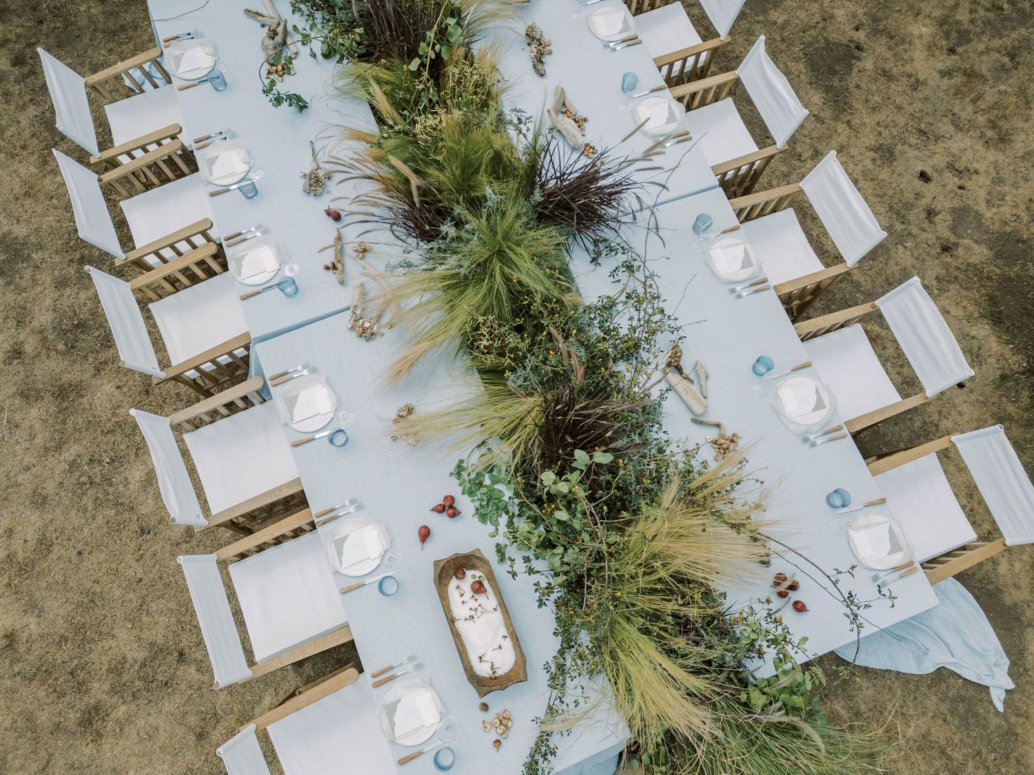 Overhead view of an elegant outdoor dining table set with white linens and natural greenery centerpieces on rustic ground.