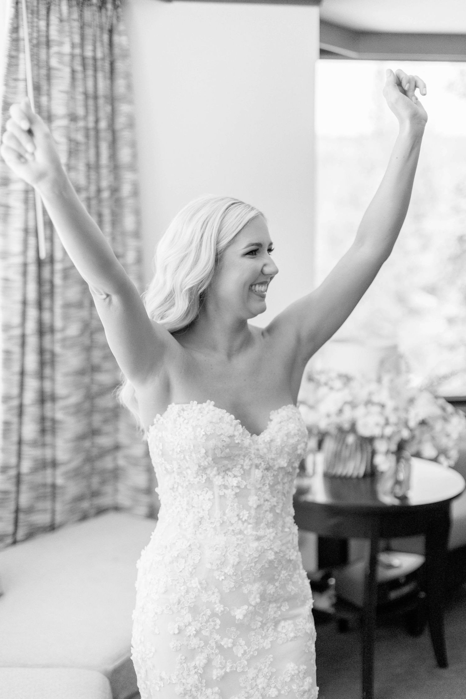 A joyful bride in a fitted lace wedding dress raises her arms in celebration.