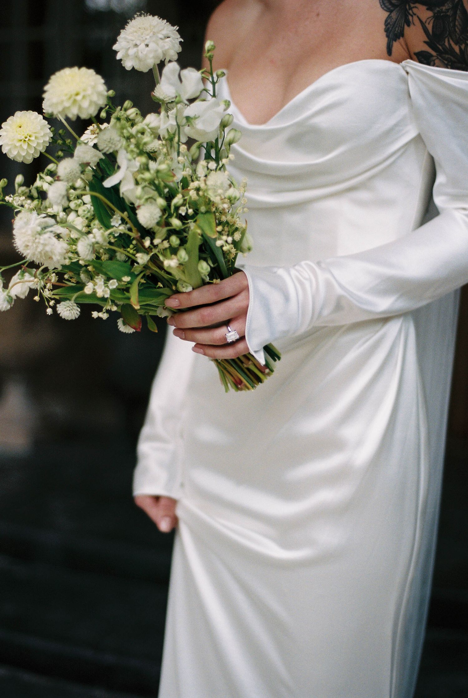 A bride in a white silk dress holds a delicate bouquet of white flowers and greenery against a dark background.