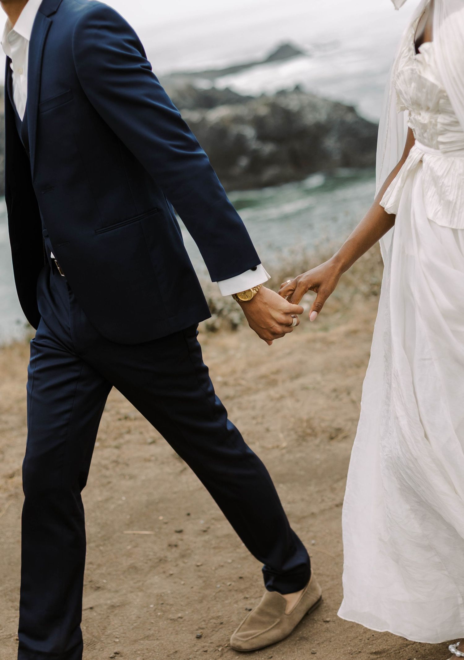 A couple in wedding attire walking along a rocky coastal shoreline holding hands in a romantic moment.