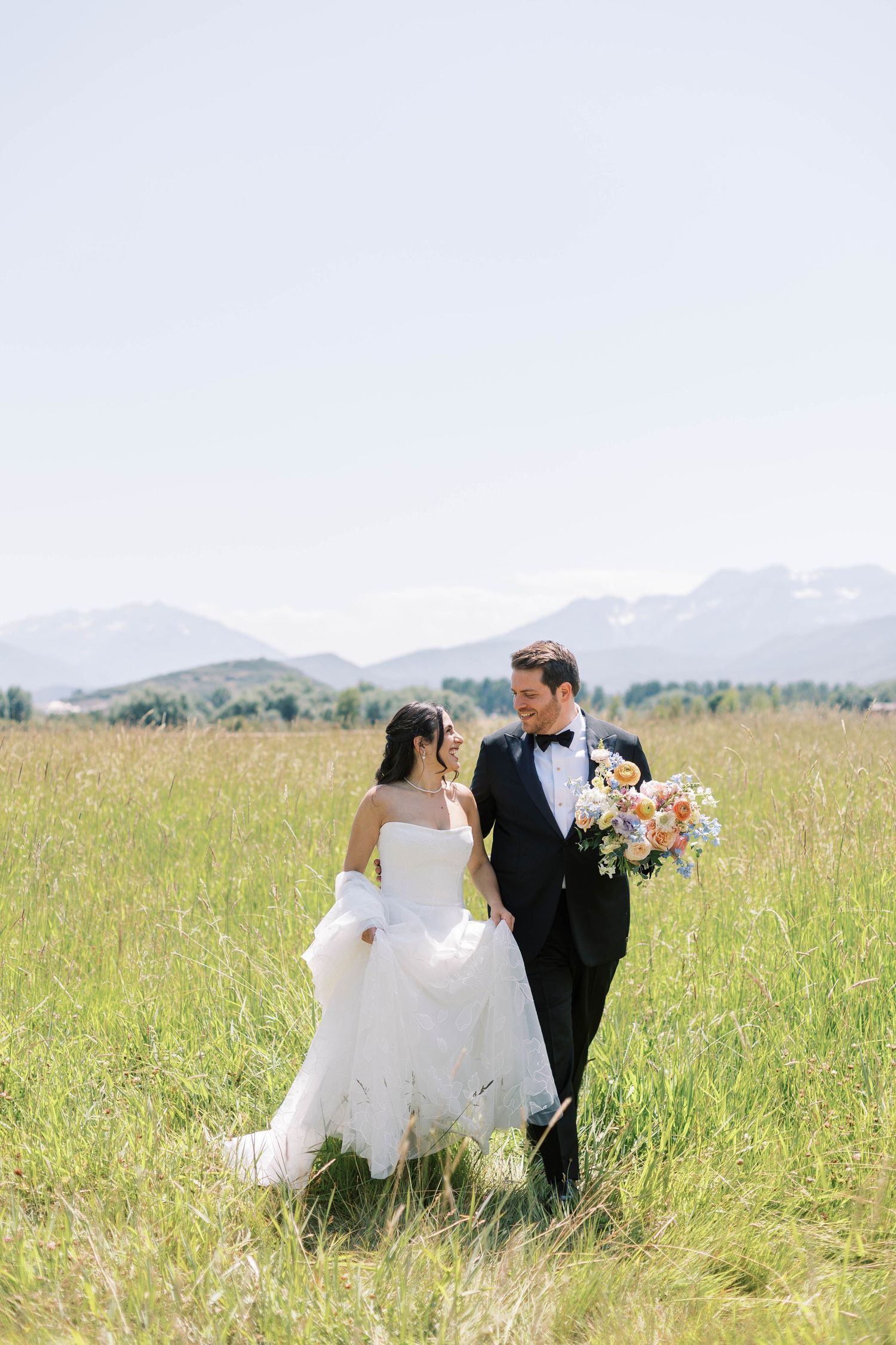 A bride and groom walking together through a green meadow with mountains in the background.