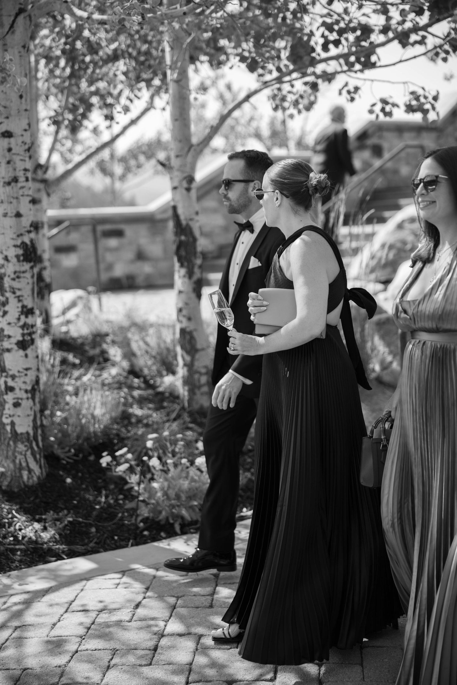 A group in formal black and gray attire stands together outdoors in a garden setting captured in black and white photography.
