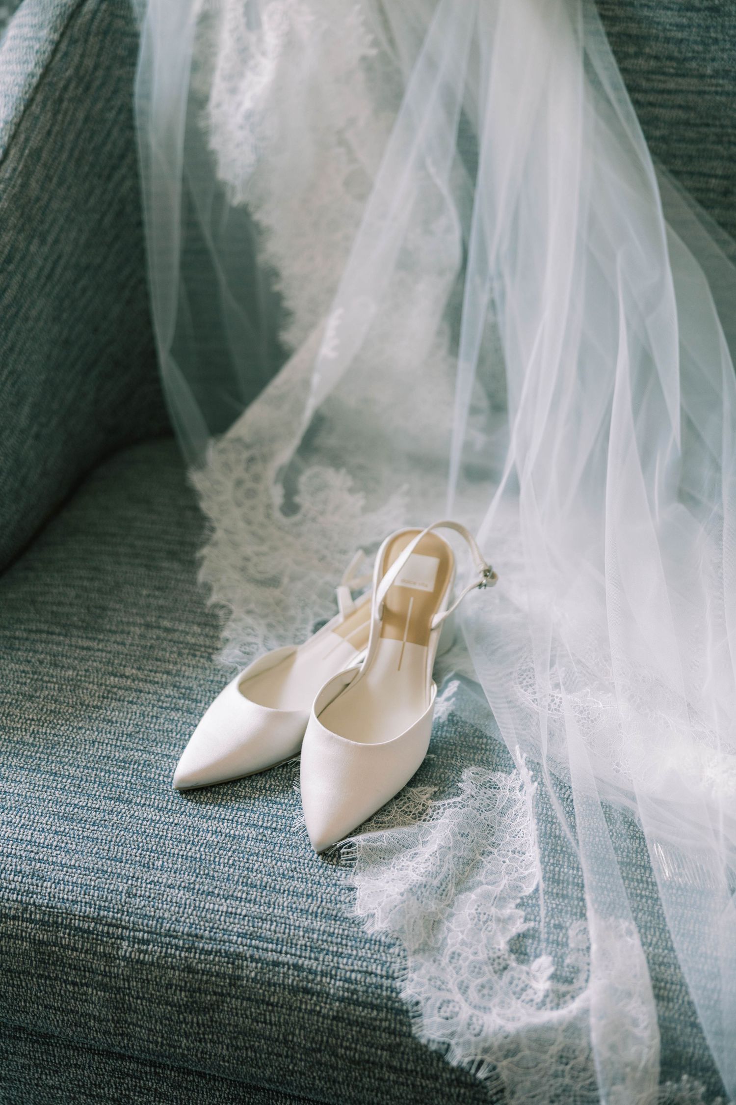 Elegant white bridal shoes with delicate lace veil draped across them on a textured gray surface.