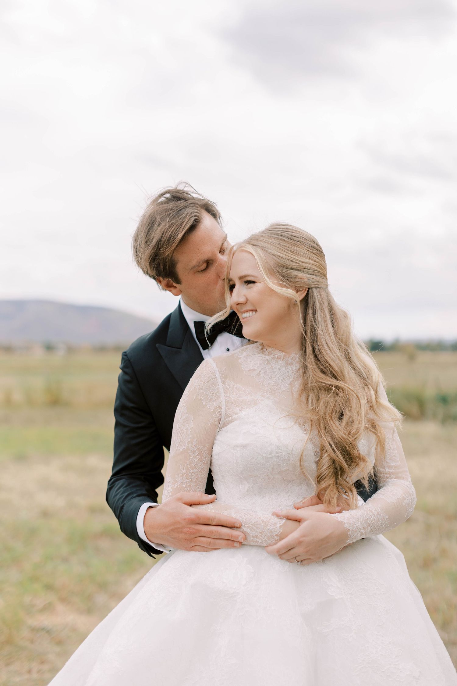 A romantic wedding couple embraces in a field under cloudy skies, the bride wearing a white lace gown.