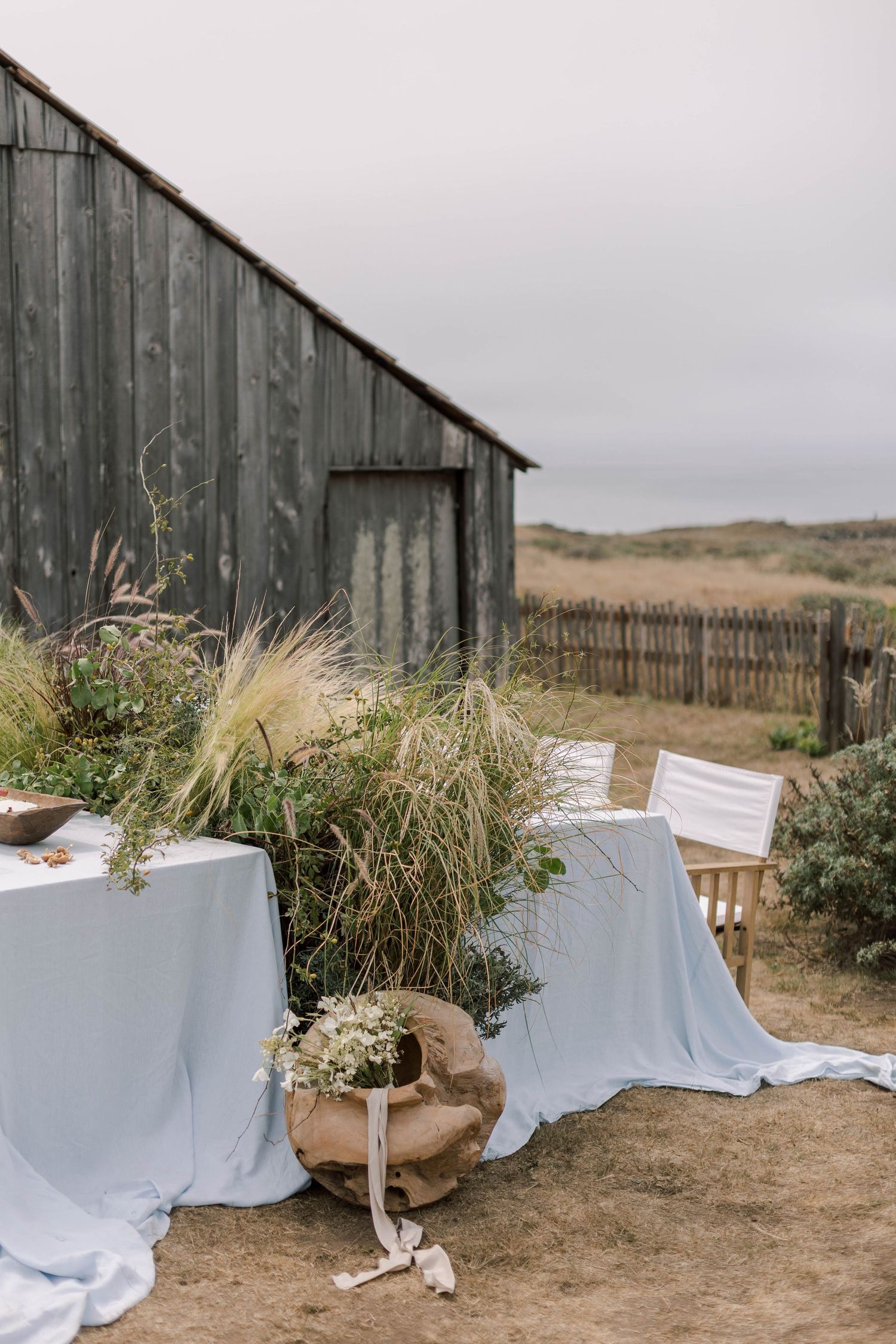 Rustic outdoor wedding setup with white tablecloths and natural grass decorations against weathered wooden barn wall.