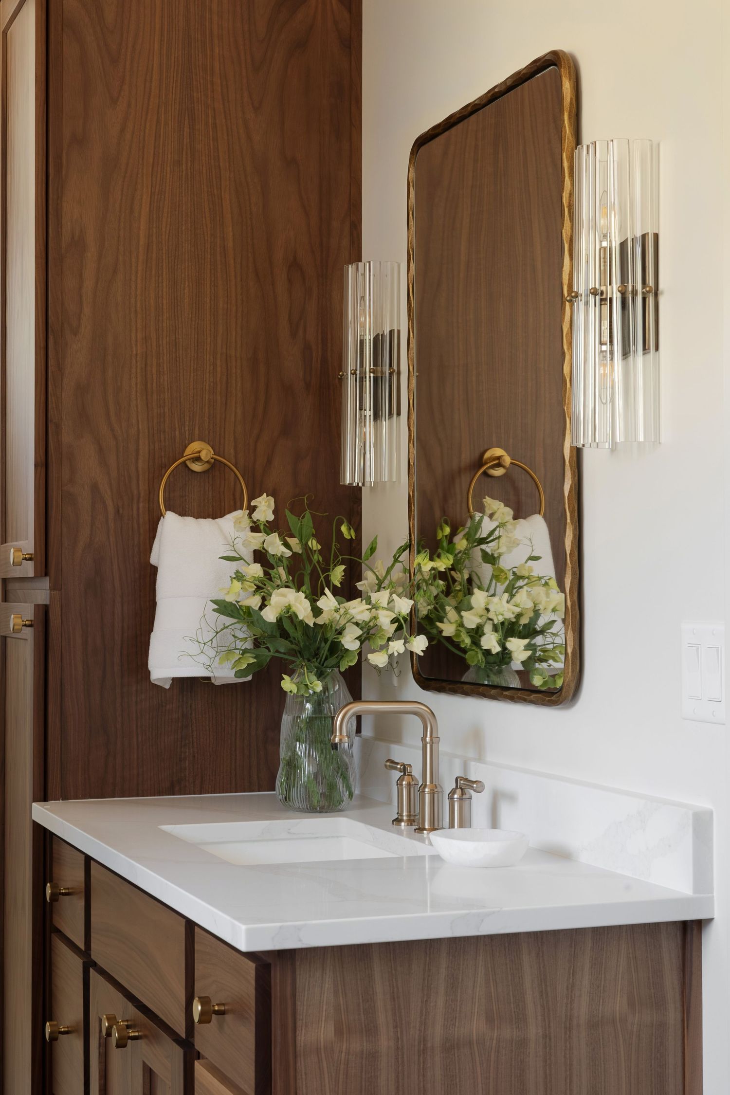 Elegant bathroom vanity with walnut cabinets, white marble countertop, brass fixtures, arched mirror, crystal sconces, and fresh white flowe