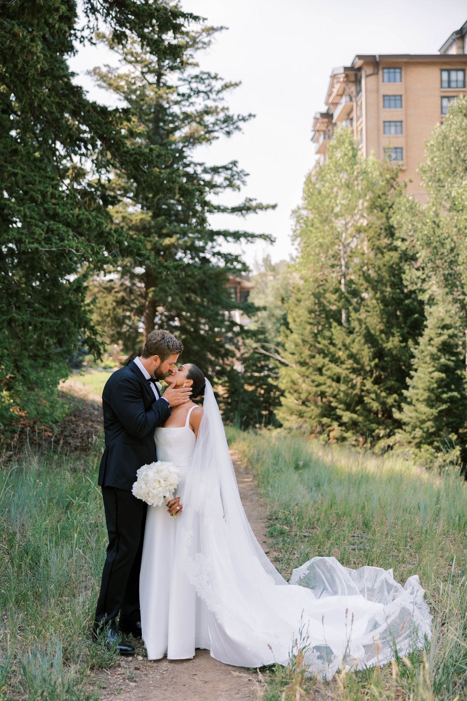 A wedding couple shares intimate moments in a scenic outdoor setting with tall grass and apartment buildings in background.