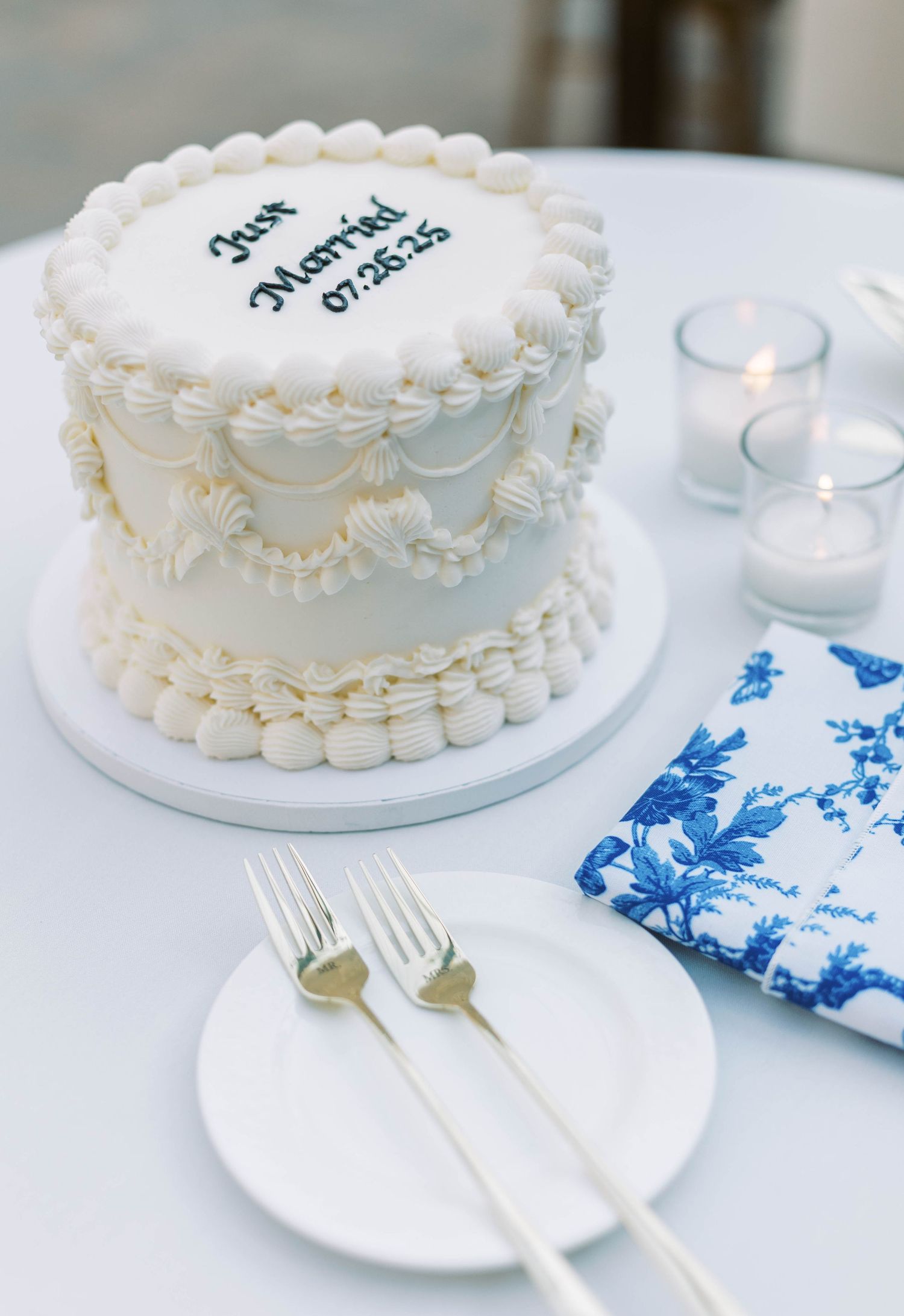 Elegant white wedding cake with delicate piping details sits beside silver forks and blue floral napkin with lit tea lights.