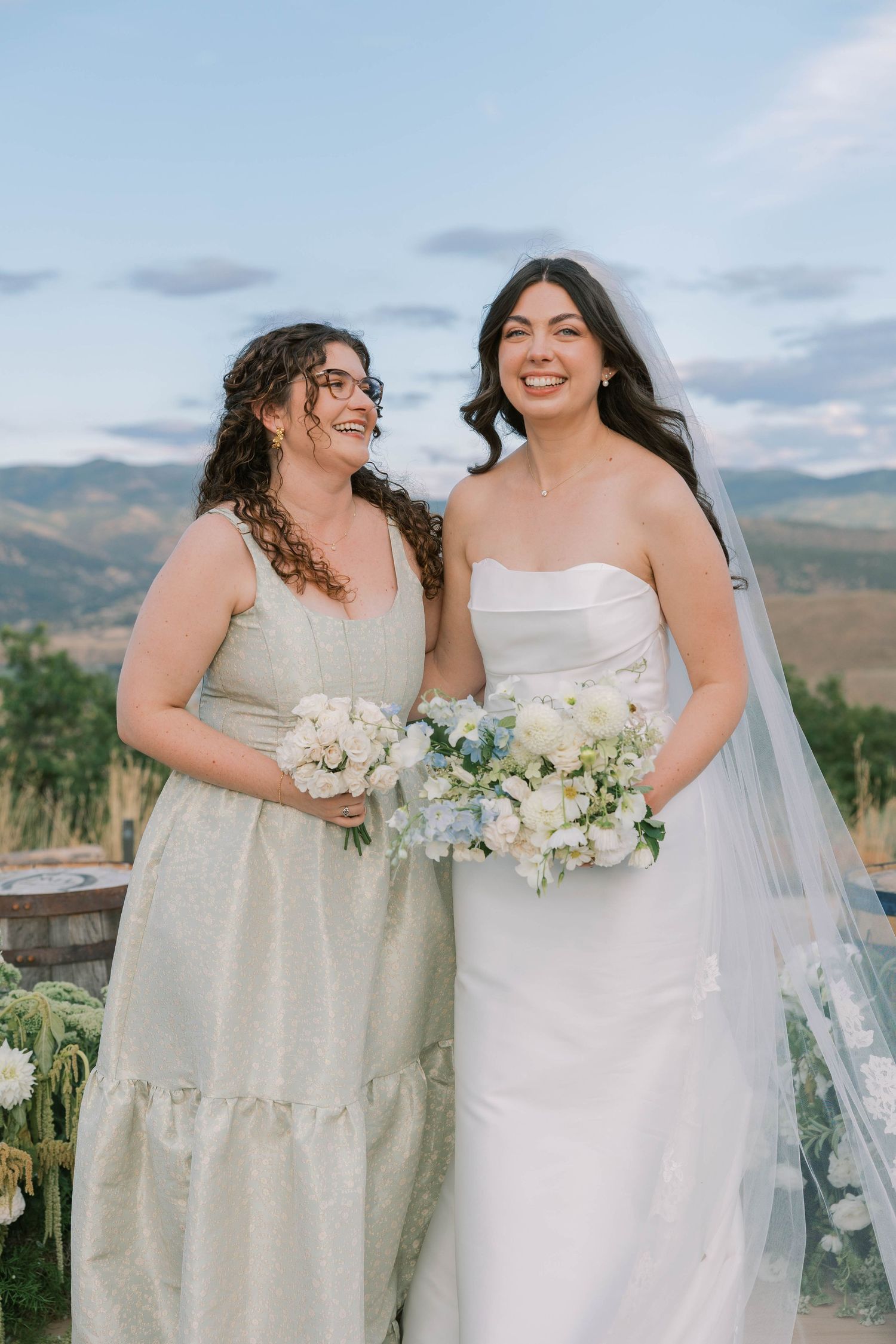 Two people in wedding attire stand together holding white floral bouquets against a scenic mountain backdrop.