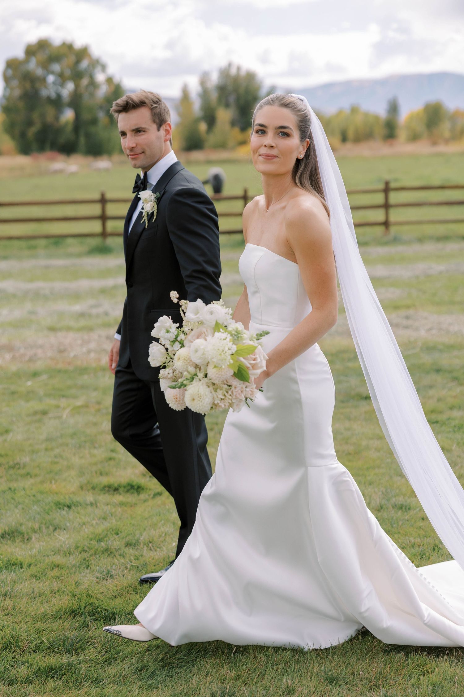 A wedding couple poses in a rustic field with mountain views as the bride holds a white floral bouquet.