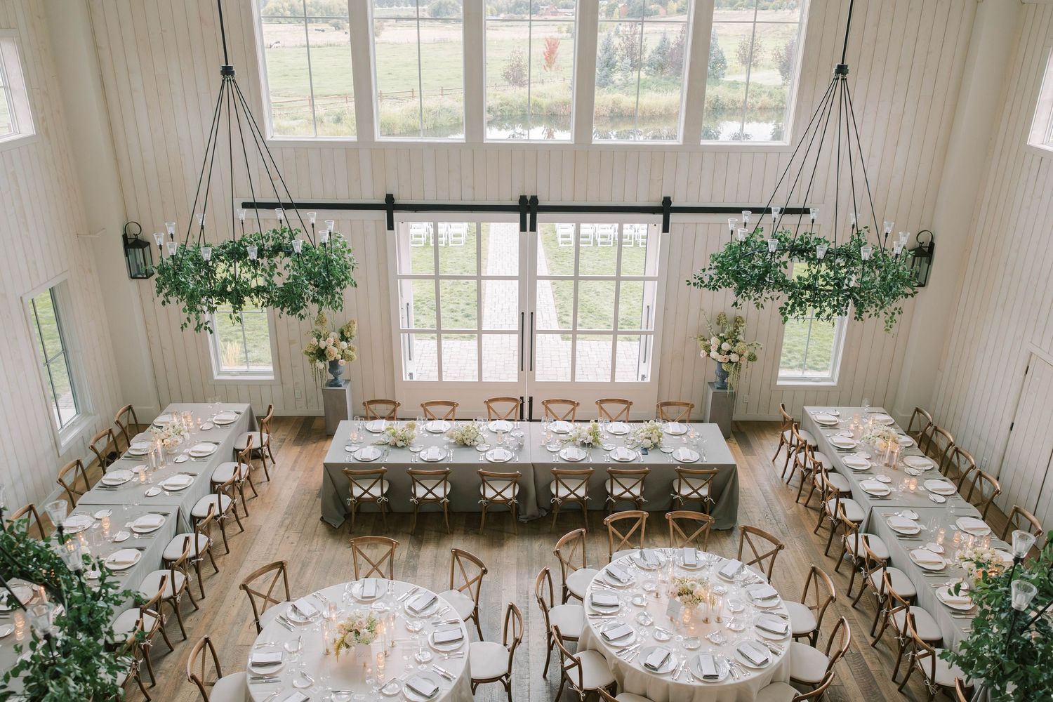 Elegant wedding reception venue with white tables, hanging greenery and natural light streaming through large windows.