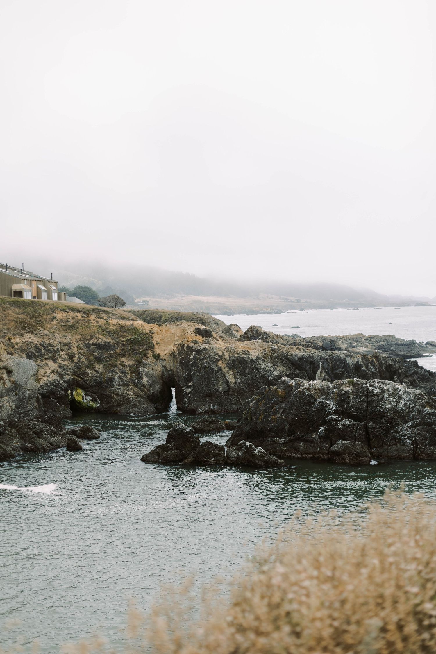 Rugged coastal landscape with rocky cliffs and crashing waves along a misty shoreline on an overcast day.