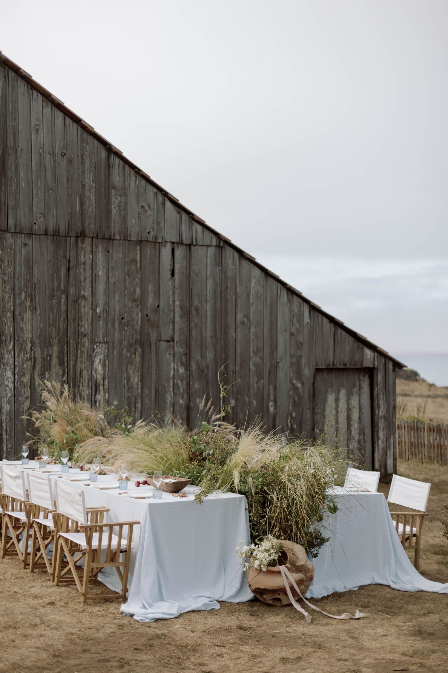Rustic outdoor wedding setup with white tablecloths and wooden chairs against a weathered barn wall in a desert setting.