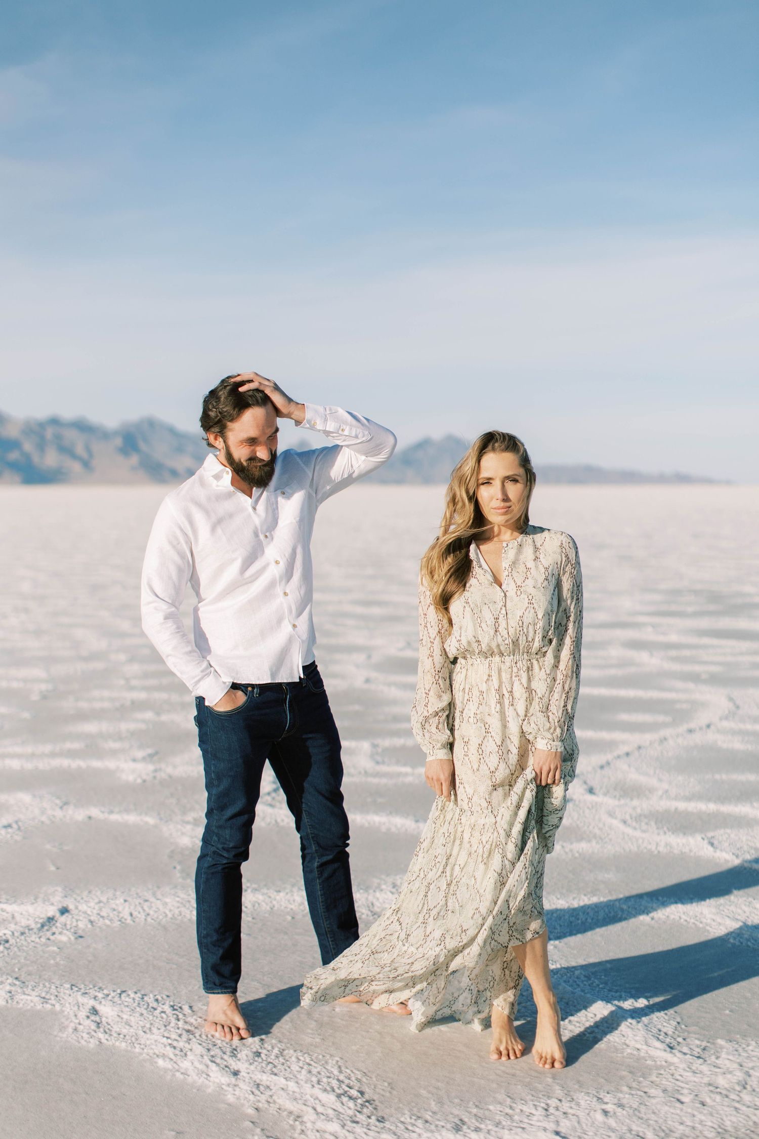 Couple in contrasting outfits posing casually on salt flats with mountains behind them.