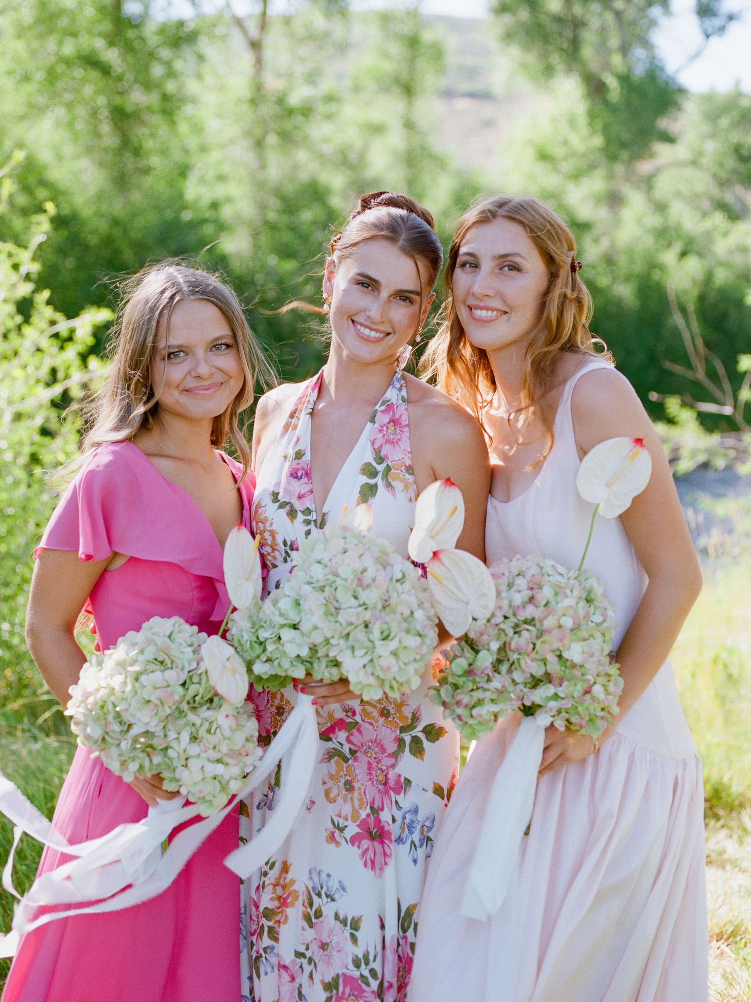 Three people in formal attire hold white and green hydrangea bouquets while posing together outdoors on a sunny day.