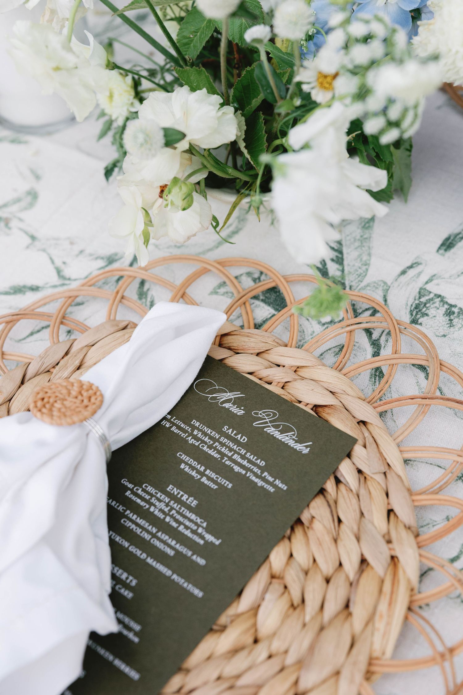 White floral arrangements with green foliage rest on woven placemats alongside dark olive green menu cards.