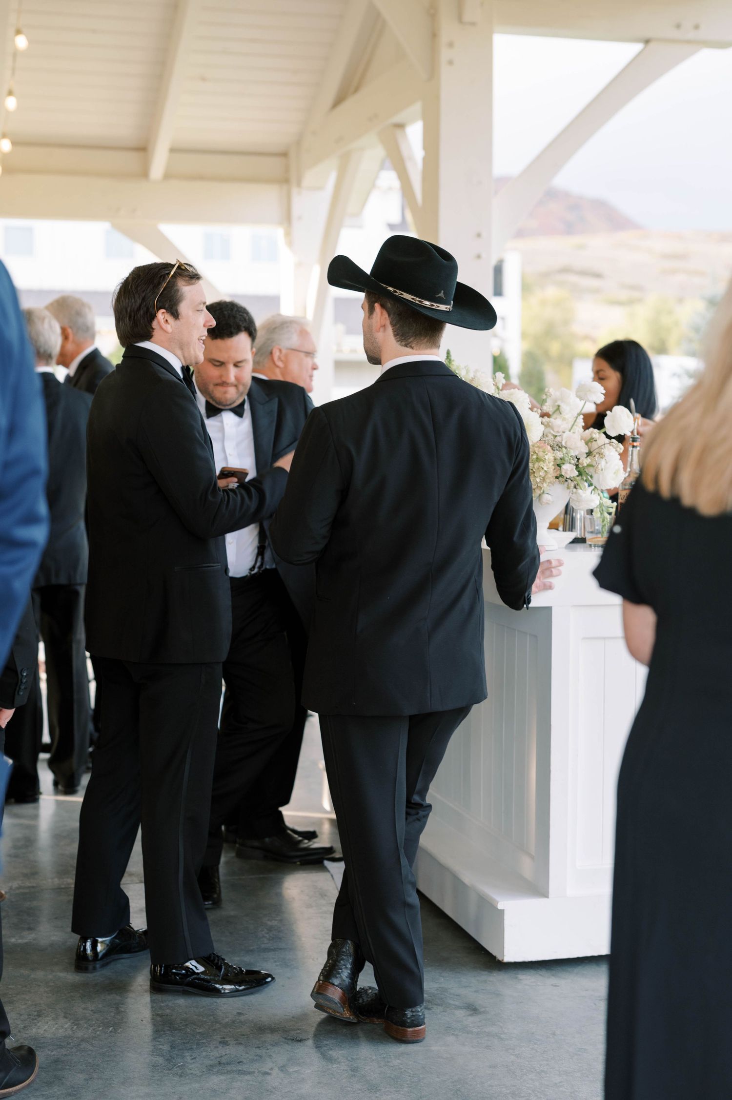 Guests in formal black attire mingle at an elegant reception under a white covered outdoor venue.
