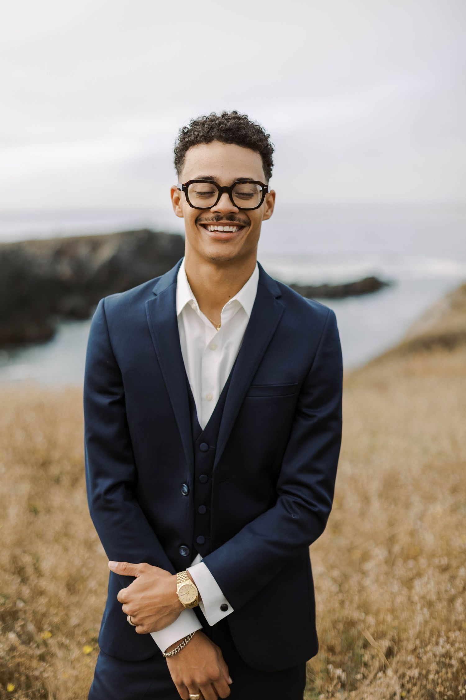 A person in a navy suit and glasses smiles warmly while standing in a golden field on an overcast day.