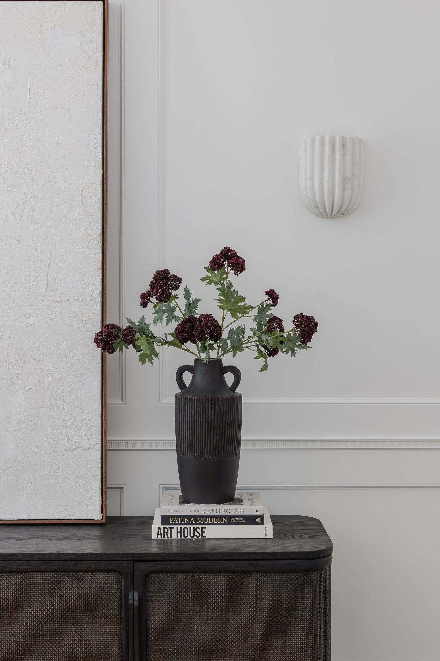 Dark textured vase with burgundy flowers on stacked books atop a rattan cabinet, beside a white wall sconce.