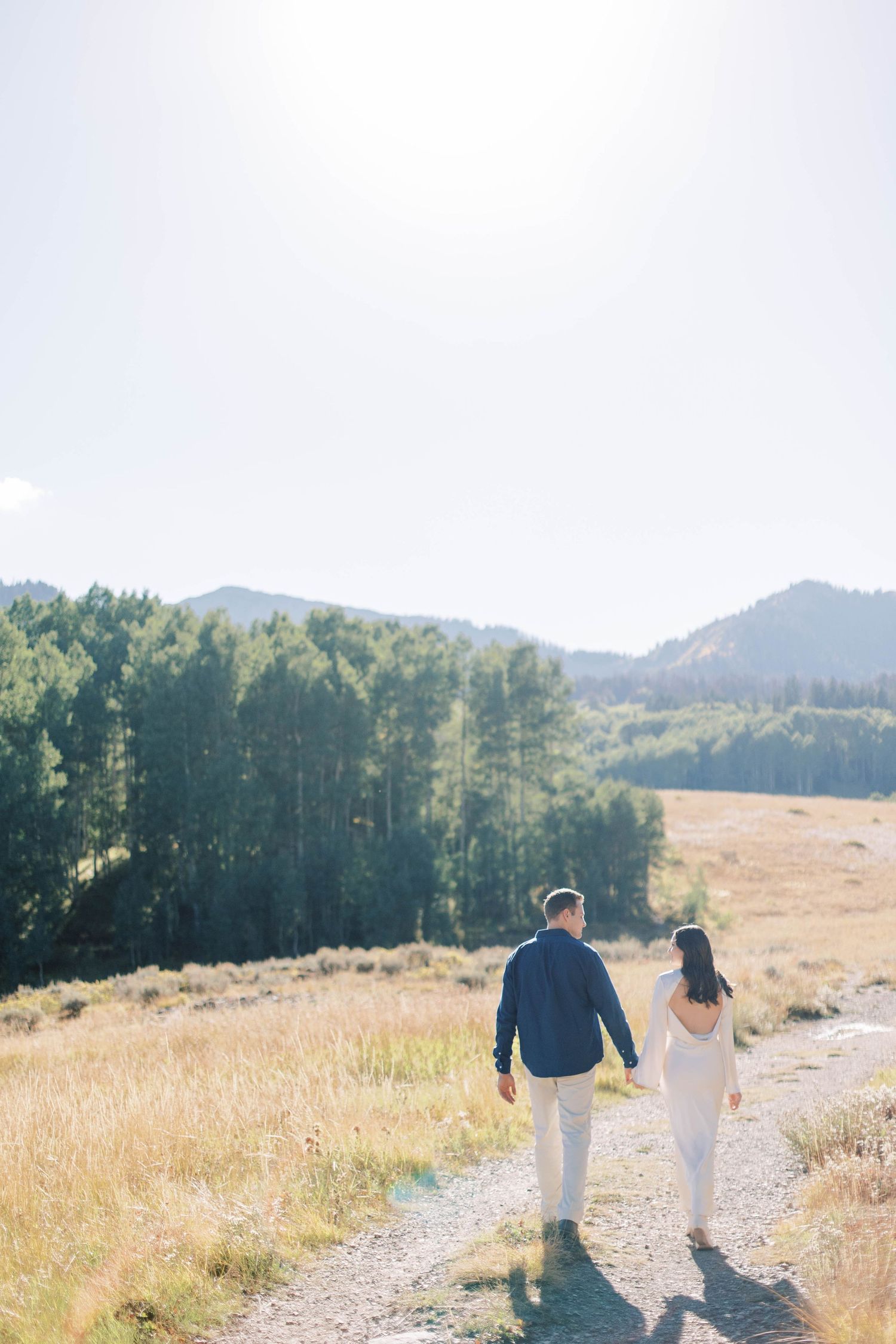 A couple walks hand in hand on a mountain trail through golden grasslands and evergreen forests.