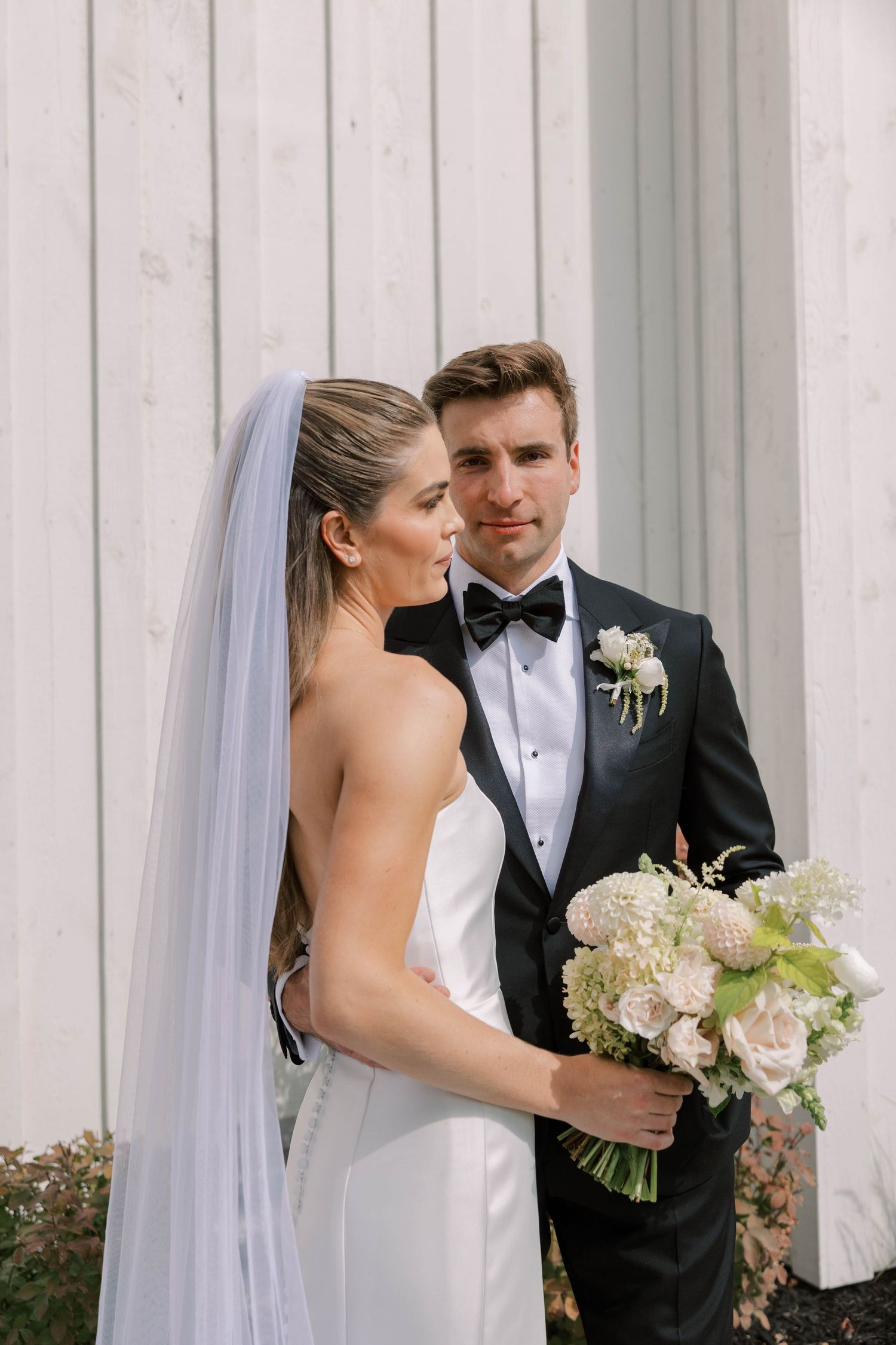 A couple in formal wedding attire standing together against a white wooden wall.