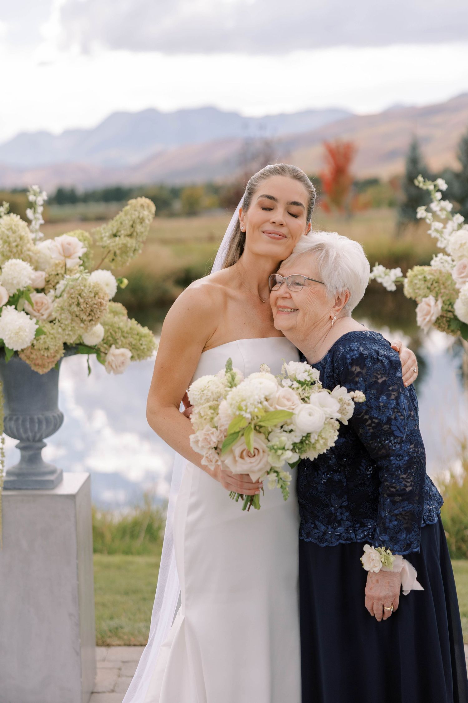 A tender moment between a bride in white and an older woman in navy lace against a mountain backdrop.