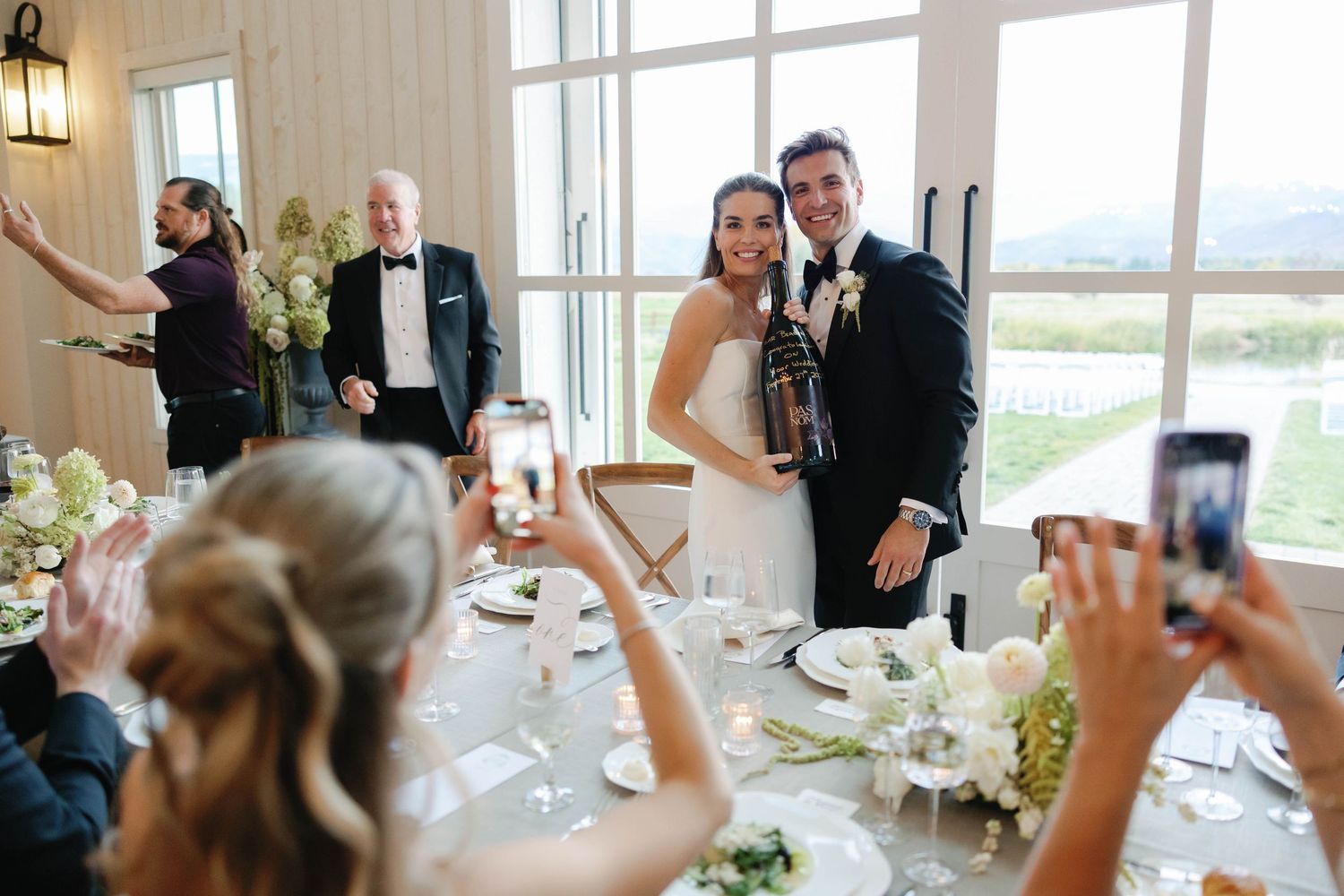Wedding guests raise their glasses in a toast during an elegant reception celebration at a bright, window-filled venue.