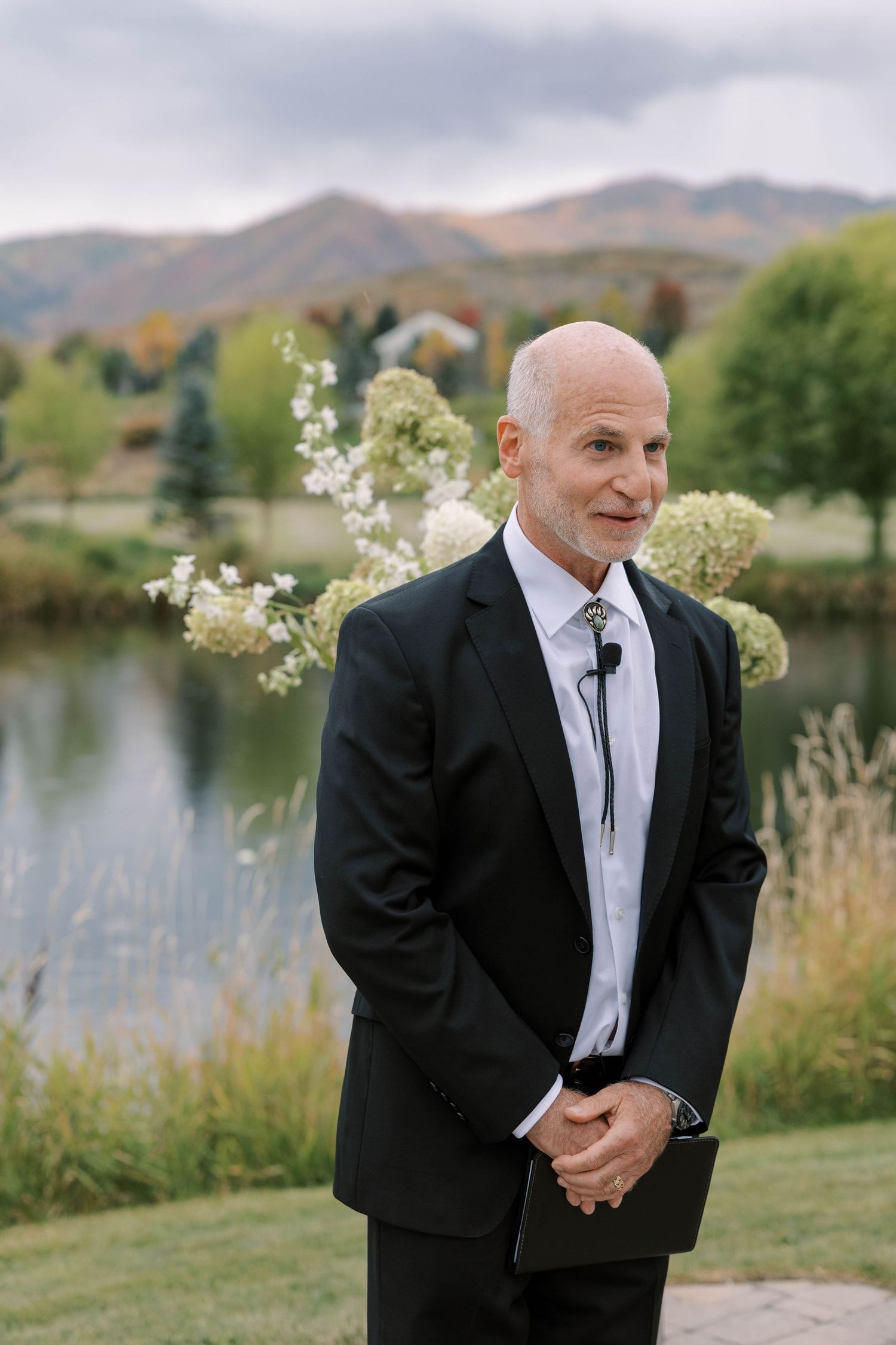 A person in a black suit and tie reacts emotionally during an outdoor wedding ceremony by a lake with mountains in background.