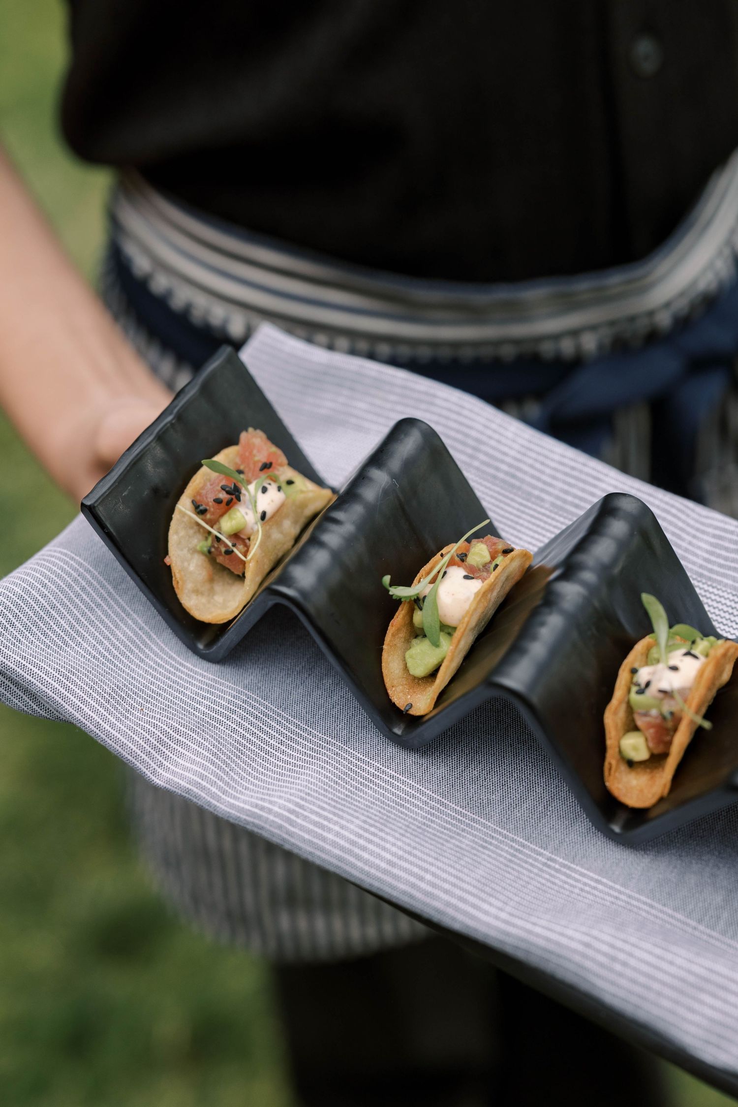 Four gourmet mini tacos served on black holders arranged on a gray napkin being presented by a server.