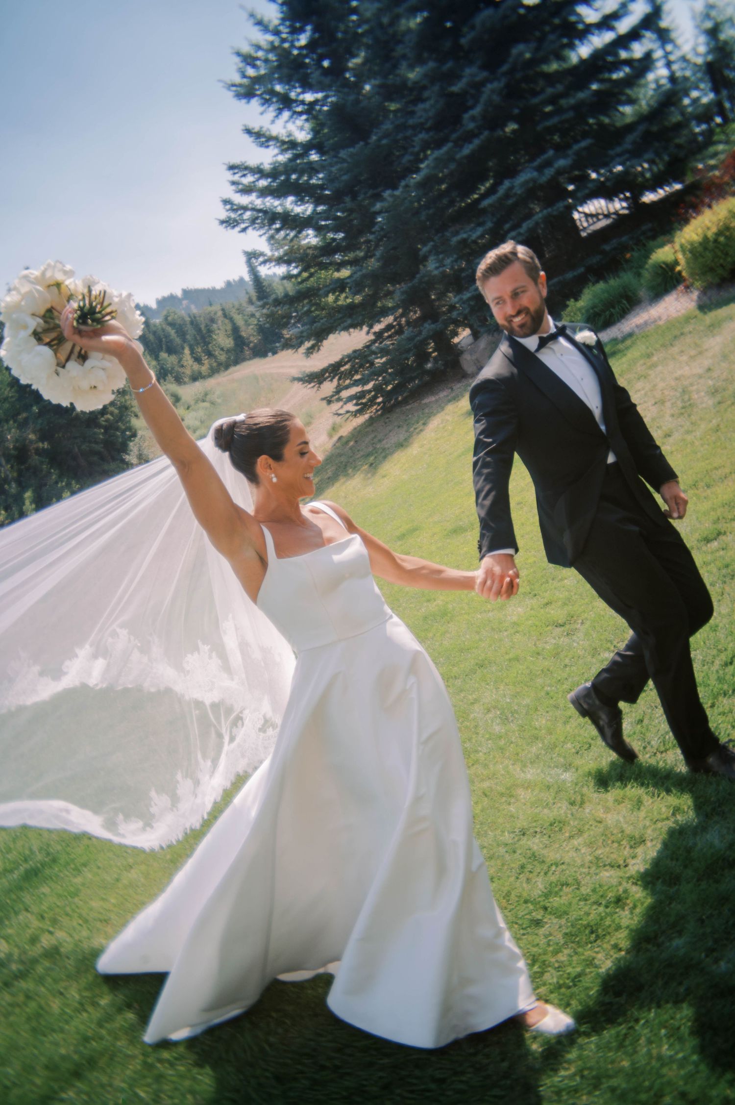 Joyful bride and groom dance playfully on a grassy hillside with evergreen trees in the background.