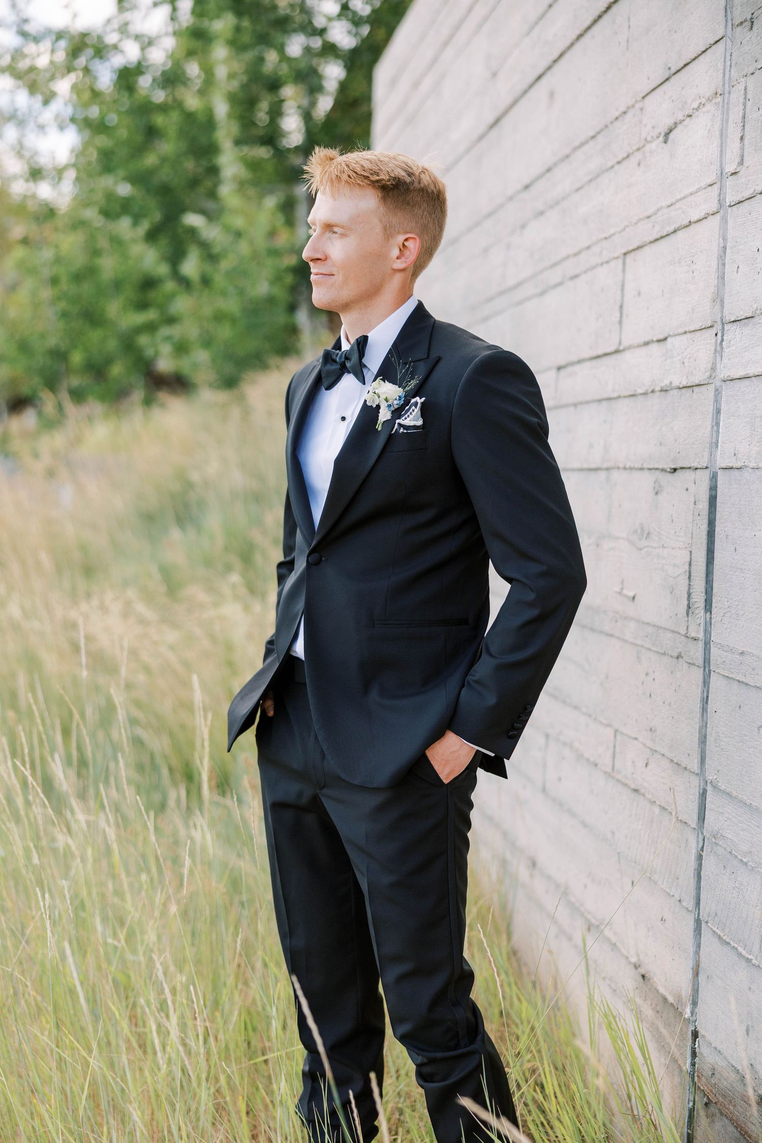 Groomsmen in black tuxedos pose against a modern concrete wall with tall grass.