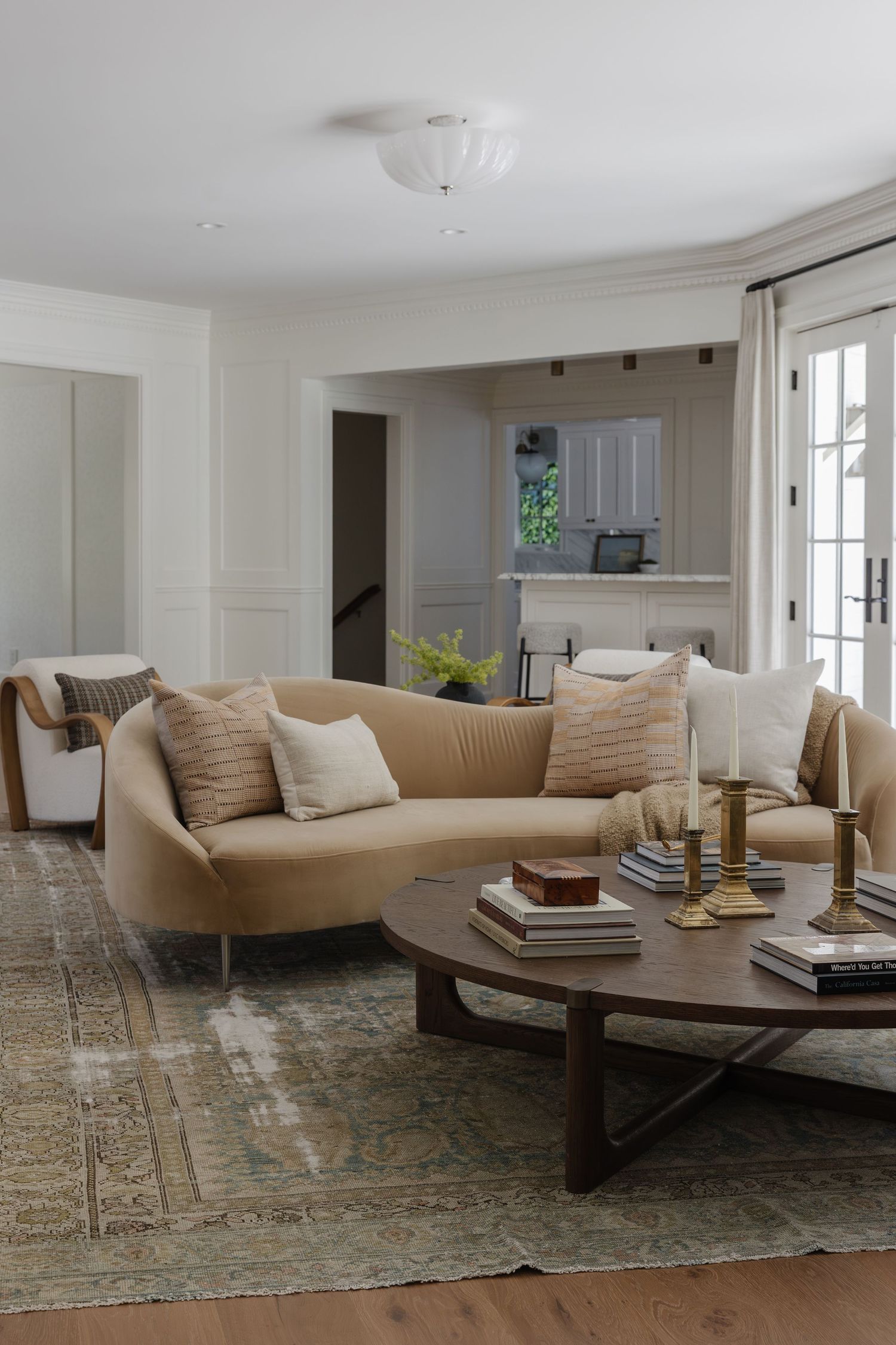 Elegant living room with curved beige sofa, round wood coffee table, vintage rug, brass candlesticks, and French doors in neutral tones.
