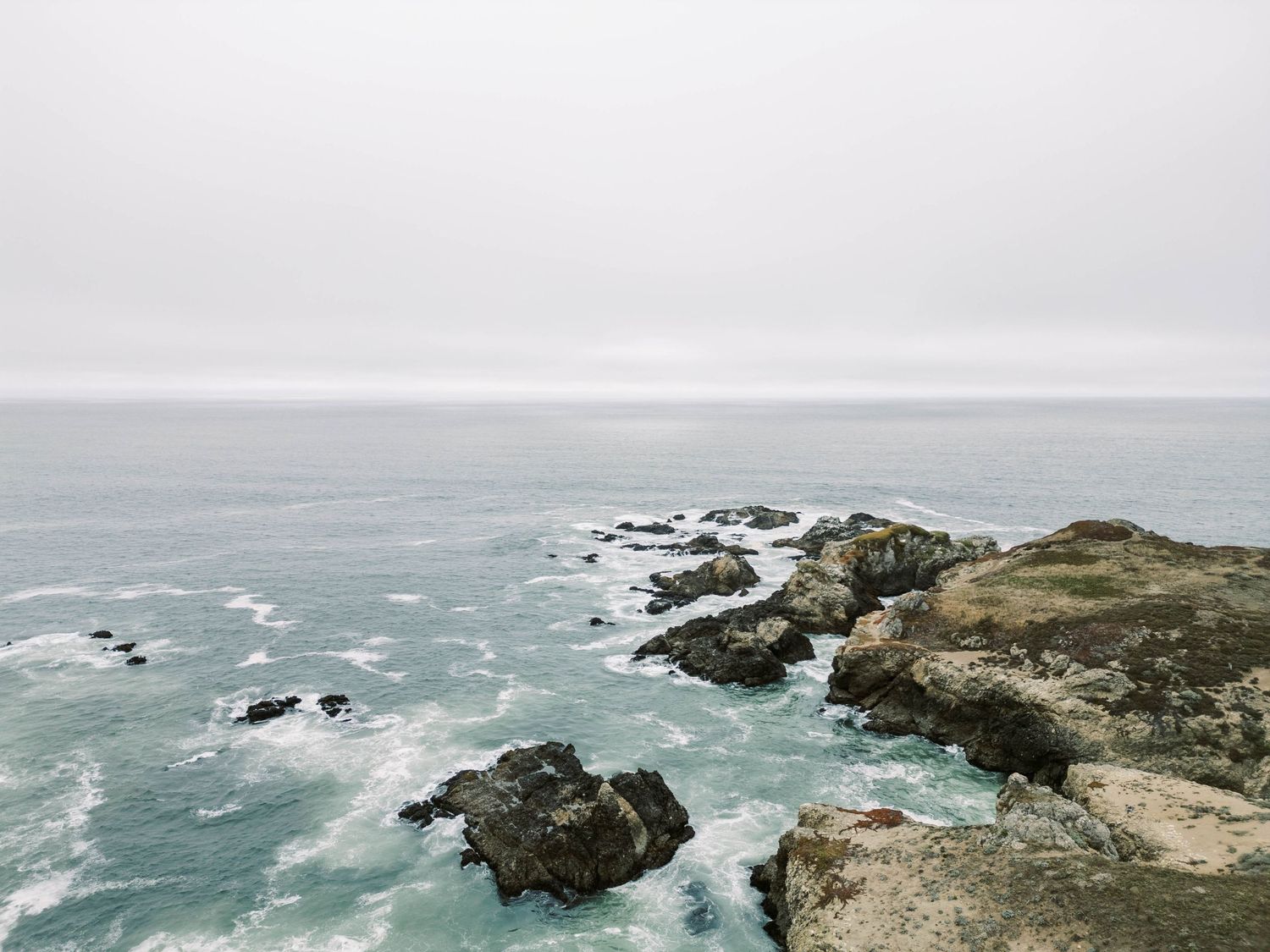 Dramatic aerial view of rugged coastal cliffs and crashing waves along a misty shoreline during overcast weather.