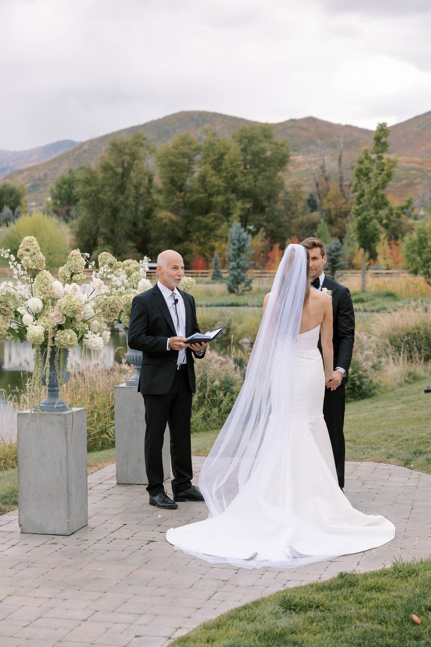 An outdoor wedding ceremony takes place on a stone patio with mountain views and floral arrangements in the background.