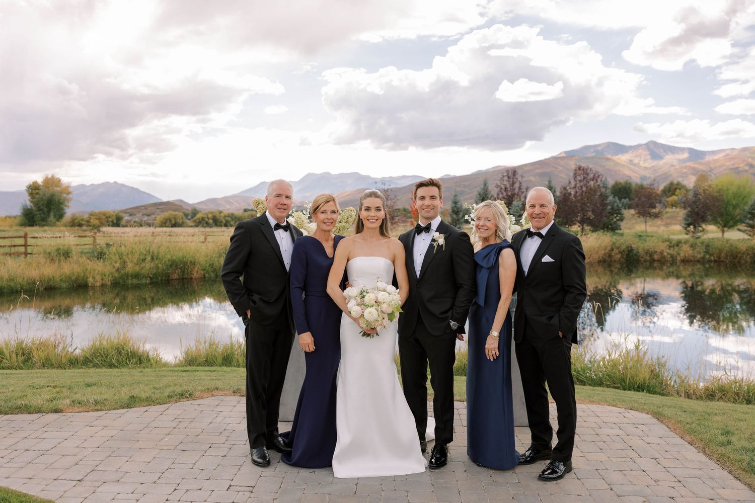 A wedding party poses outdoors in formal attire against a scenic mountain backdrop with a tranquil pond nearby.