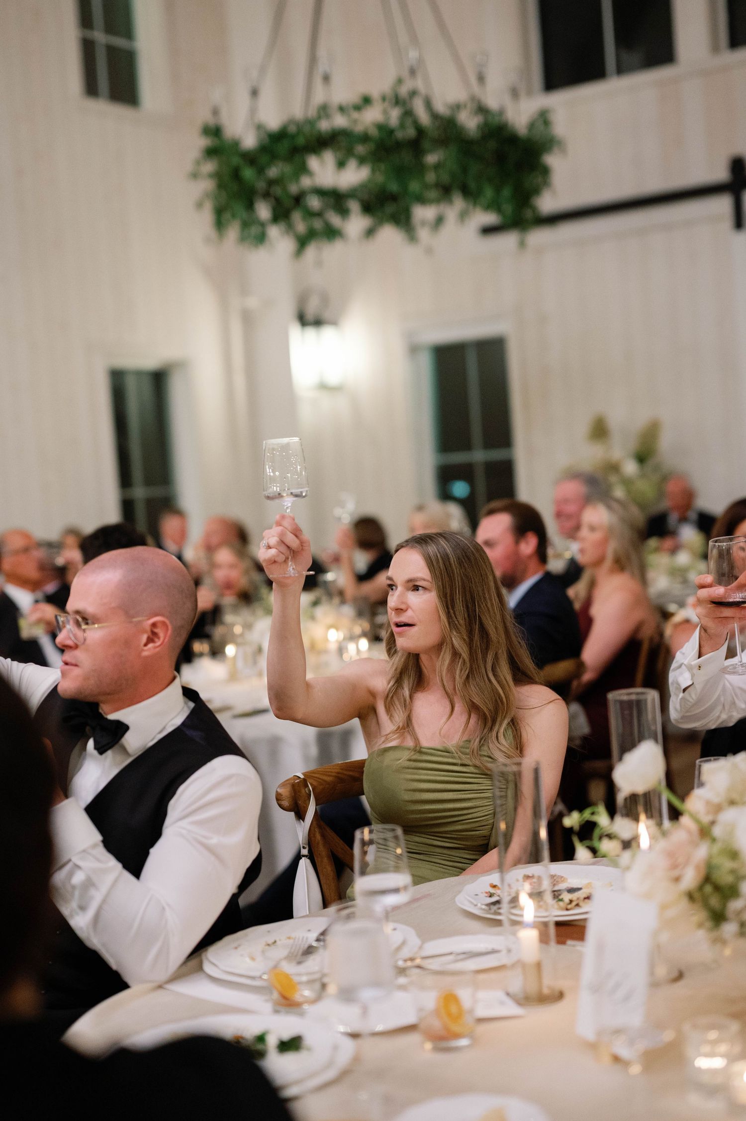 Wedding guests raise their glasses in a toast during an elegant reception dinner at a white-walled venue.